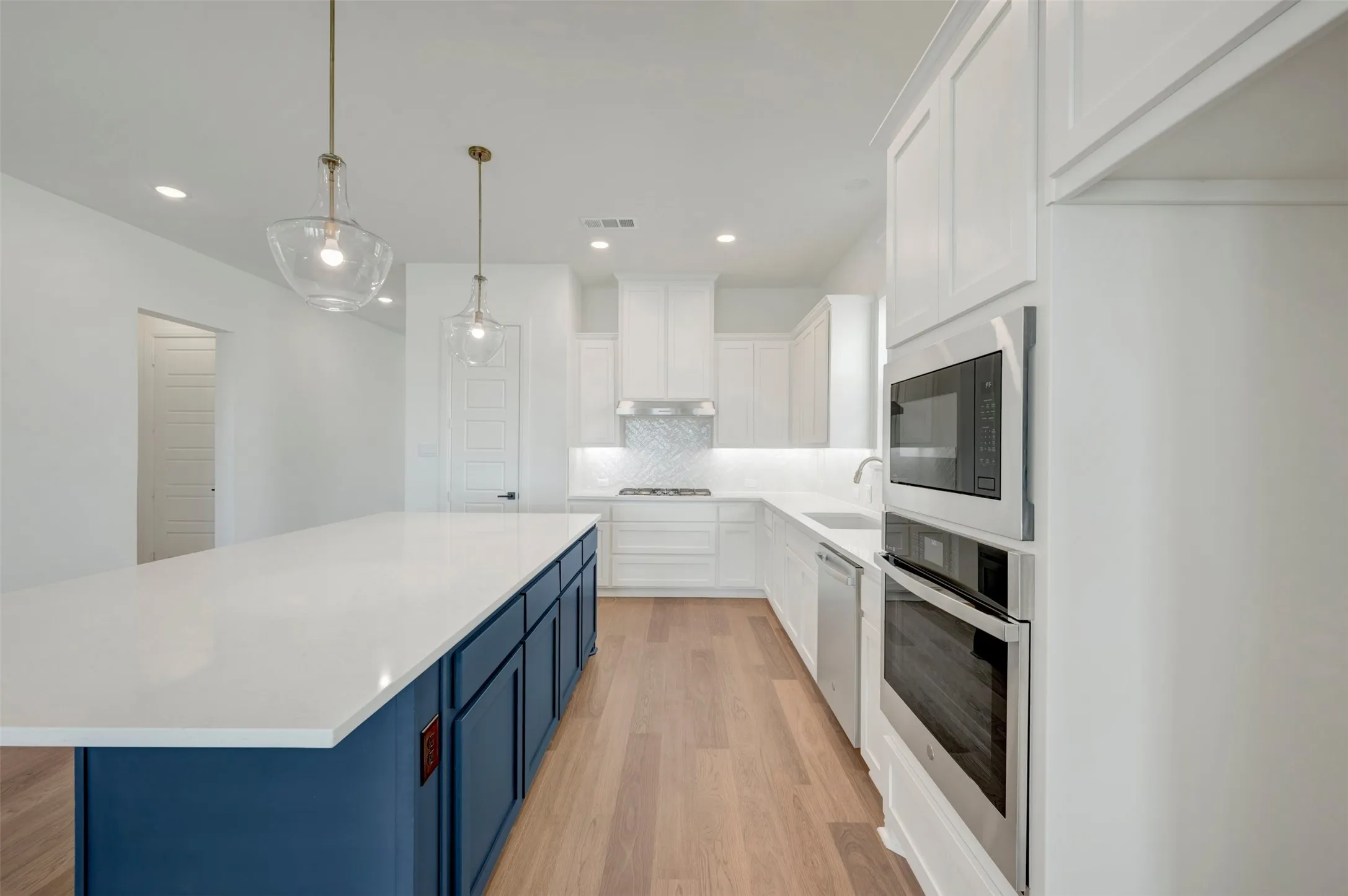 Kitchen featuring a large island with a white countertop and blue cabinetry, wood-finish flooring, white upper and lower cabinetry, stainless steel appliances, and two pendant light fixtures