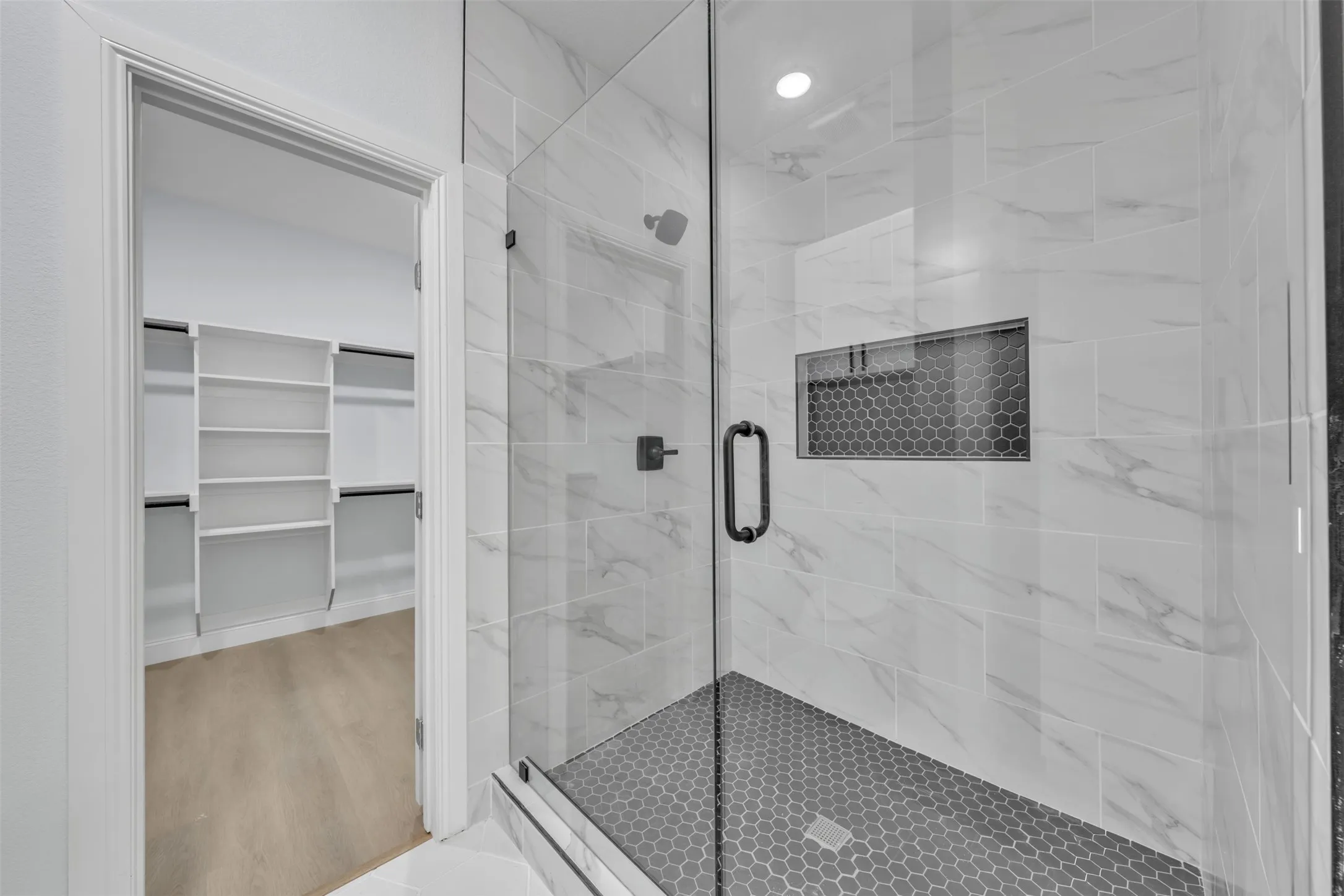 Glass-enclosed shower featuring large format white marble-look wall tiles, a dark hex-tile shower pan, and a built-in shower niche