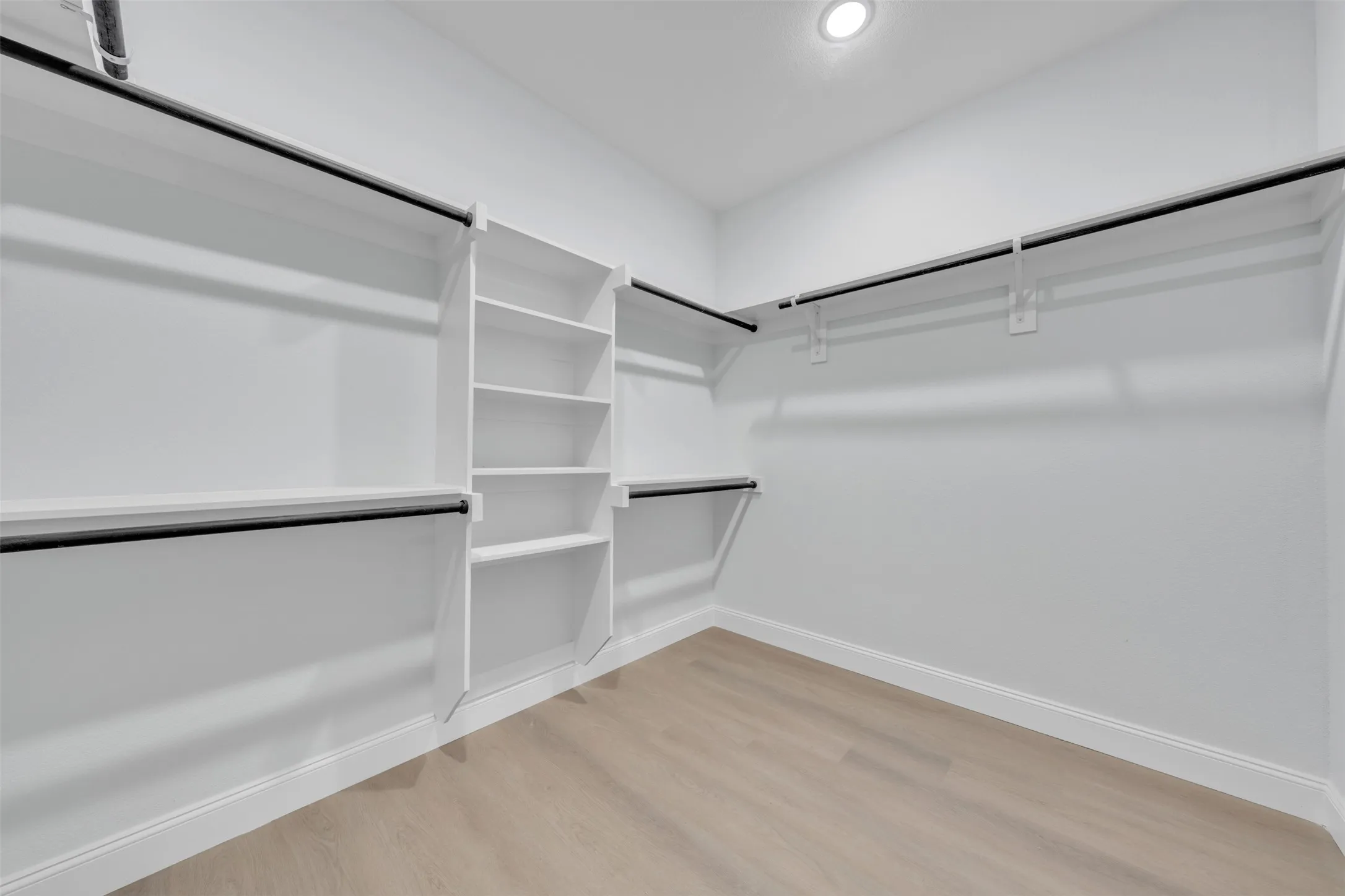 Spacious walk-in closet featuring white built-in shelving, multiple hanging rods, recessed ceiling lighting, and light wood-finish flooring