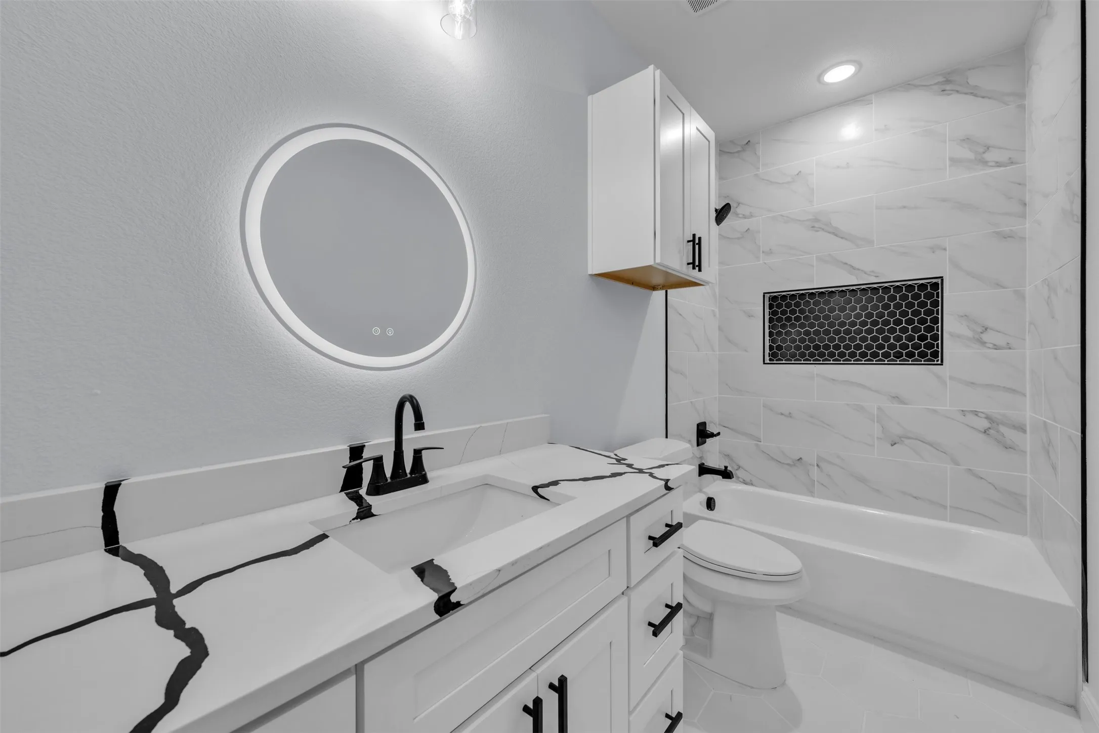 Bathroom featuring a vanity with a white countertop and black veining, a round backlit mirror, and a white soaking tub with tiled surround
