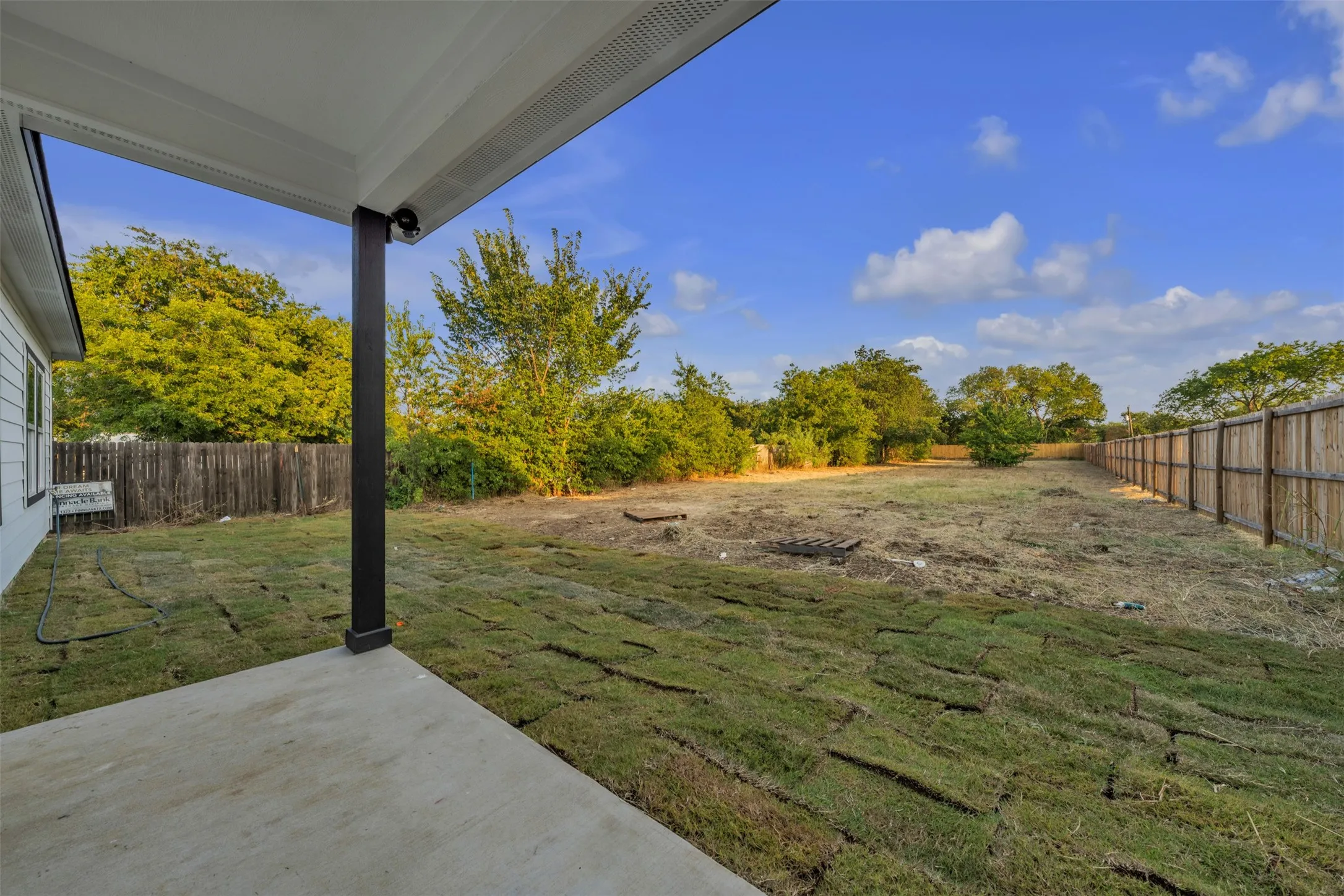 Expansive backyard featuring a concrete patio, partial sod installation, a wooden privacy fence, and mature perimeter trees