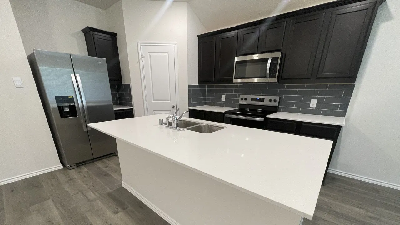 Kitchen featuring a large central island with a double basin sink, stainless steel appliances, dark wood-finish cabinetry, white countertops, and a gray subway tile backsplash