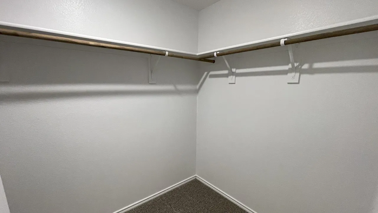Walk-in closet featuring white walls, wood-finish hanging rods, white shelving supports, and a carpeted floor