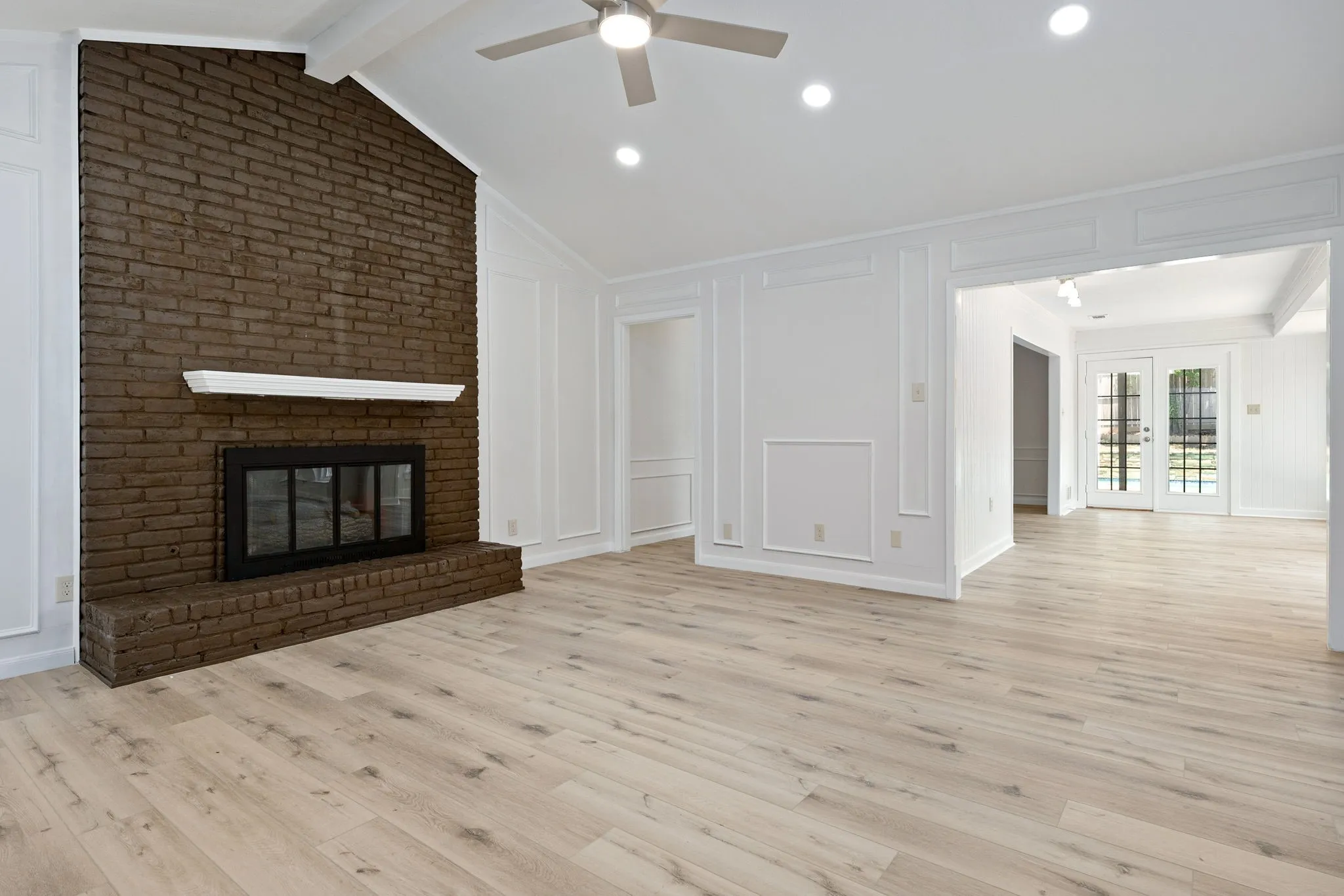 Spacious living area featuring a prominent brick fireplace with a white mantel, vaulted ceilings with exposed beams, recessed lighting, and wood-finish flooring