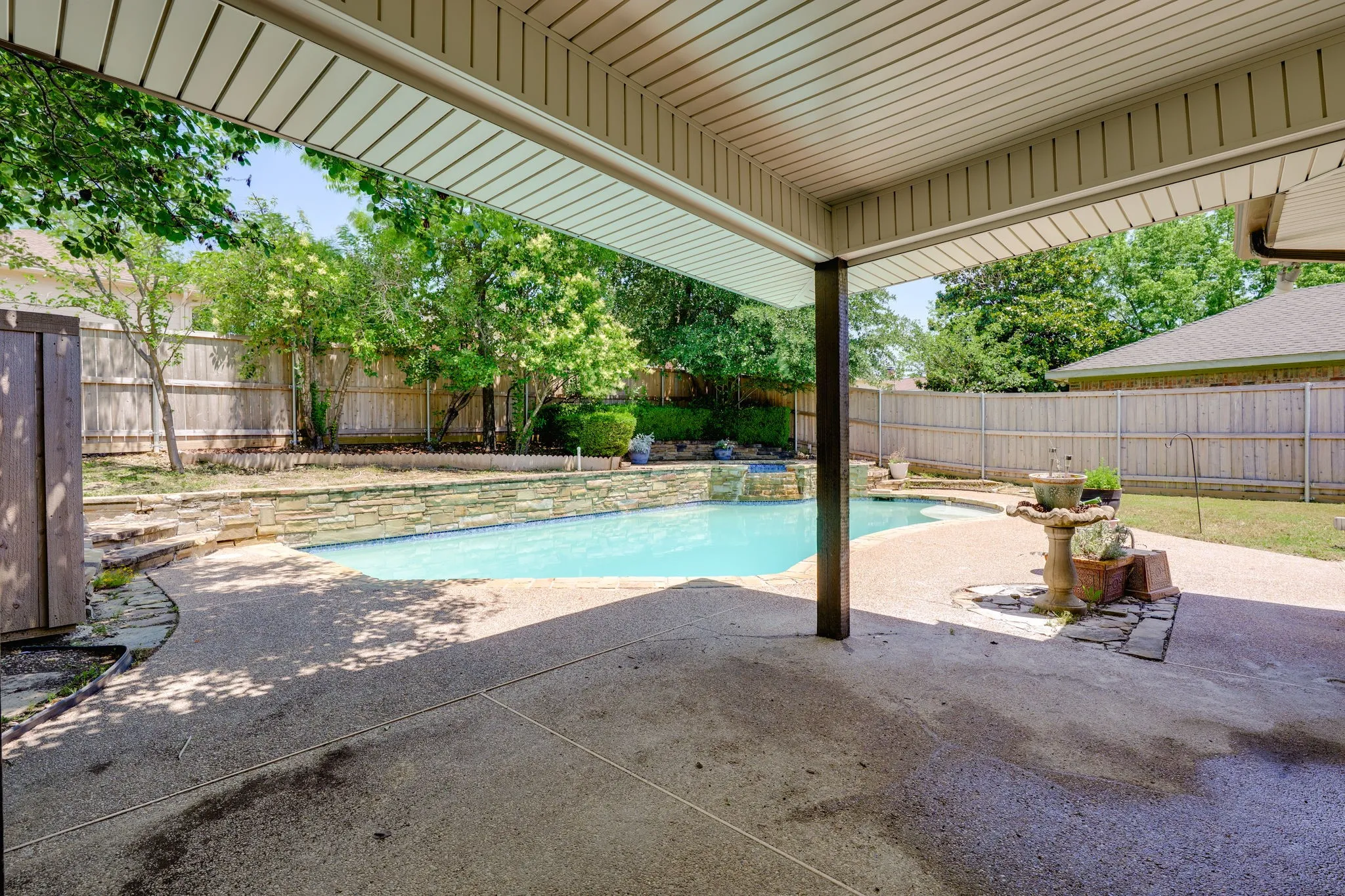 Covered patio featuring an exposed aggregate concrete surface, leading to an inground pool with a stone coping and a waterfall feature