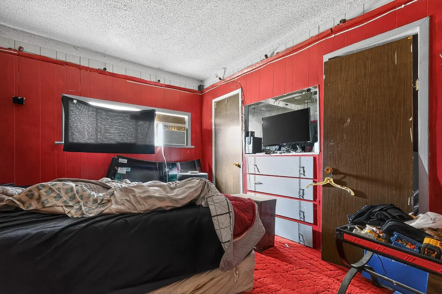Detached single bedroom featuring vertical wood paneling painted in a bold red color palette, a wall-mounted television alcove layout, built-in dresser drawers, and carpet flooring.