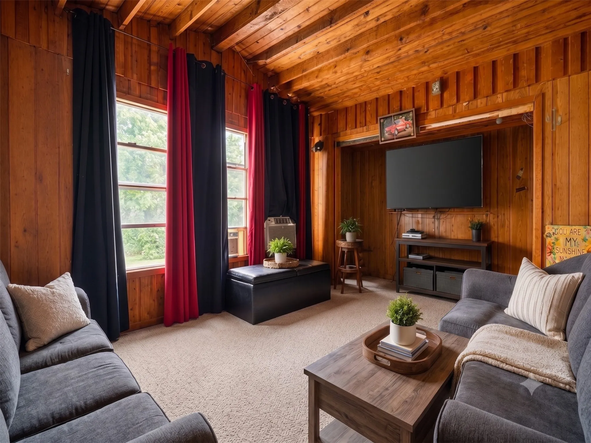 Living room area featuring natural vertical wood paneling on the walls, an exposed wood beam ceiling layout, carpet flooring, a large wall-mounted television, and multiple front-facing windows.