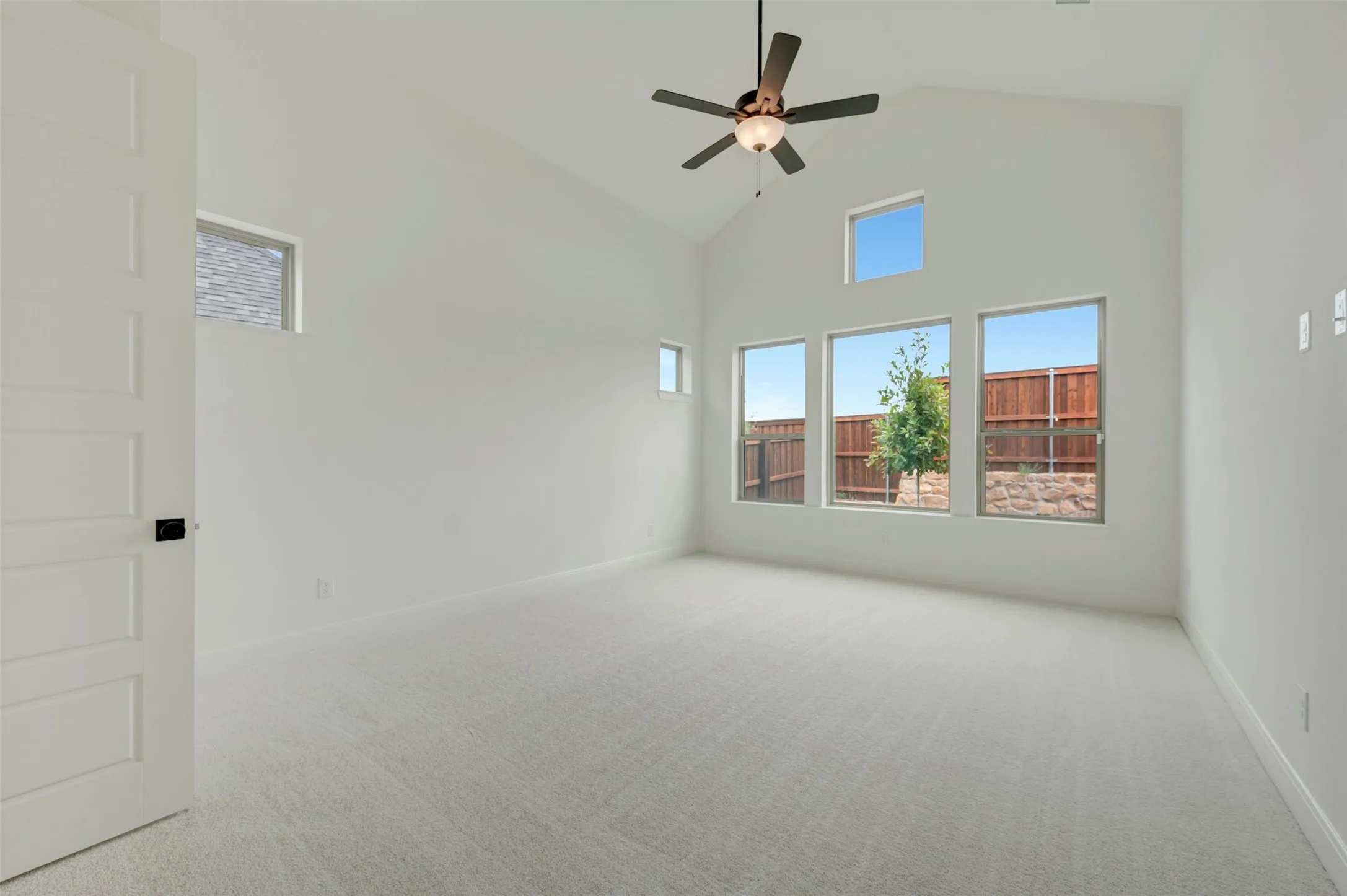 Vaulted ceiling, multiple windows, ceiling fan, neutral-toned carpet, and a paneled door
