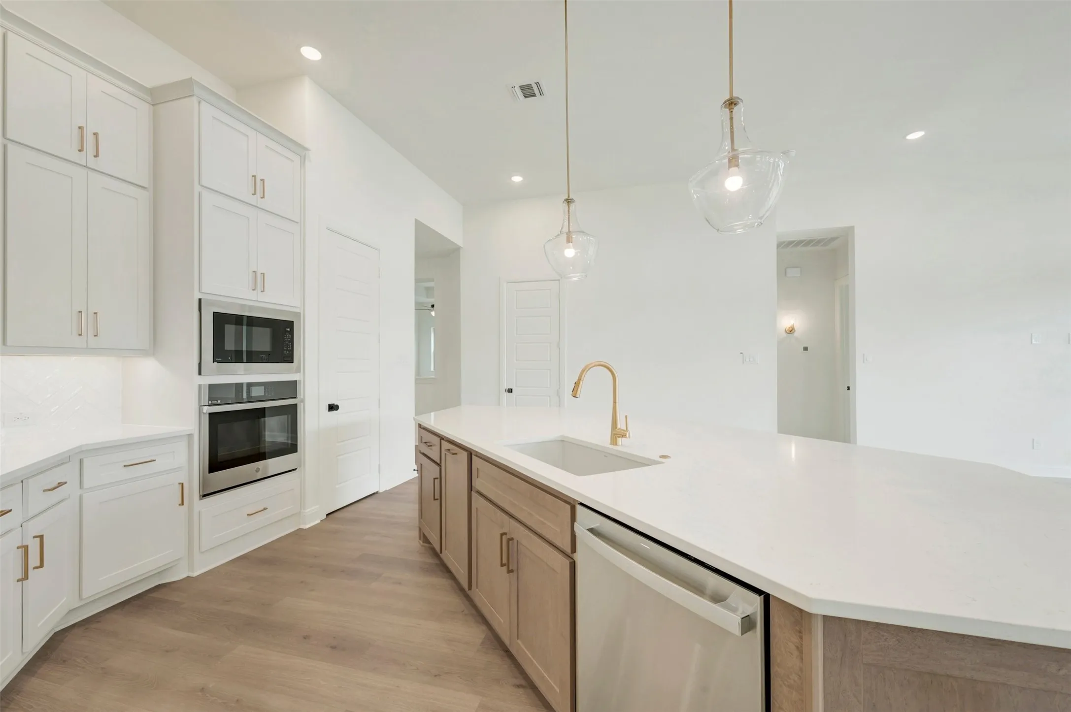 White cabinetry with gold-finish hardware, built-in stainless steel appliances, wood-finish flooring, and a large central island with a sink and pendant lighting