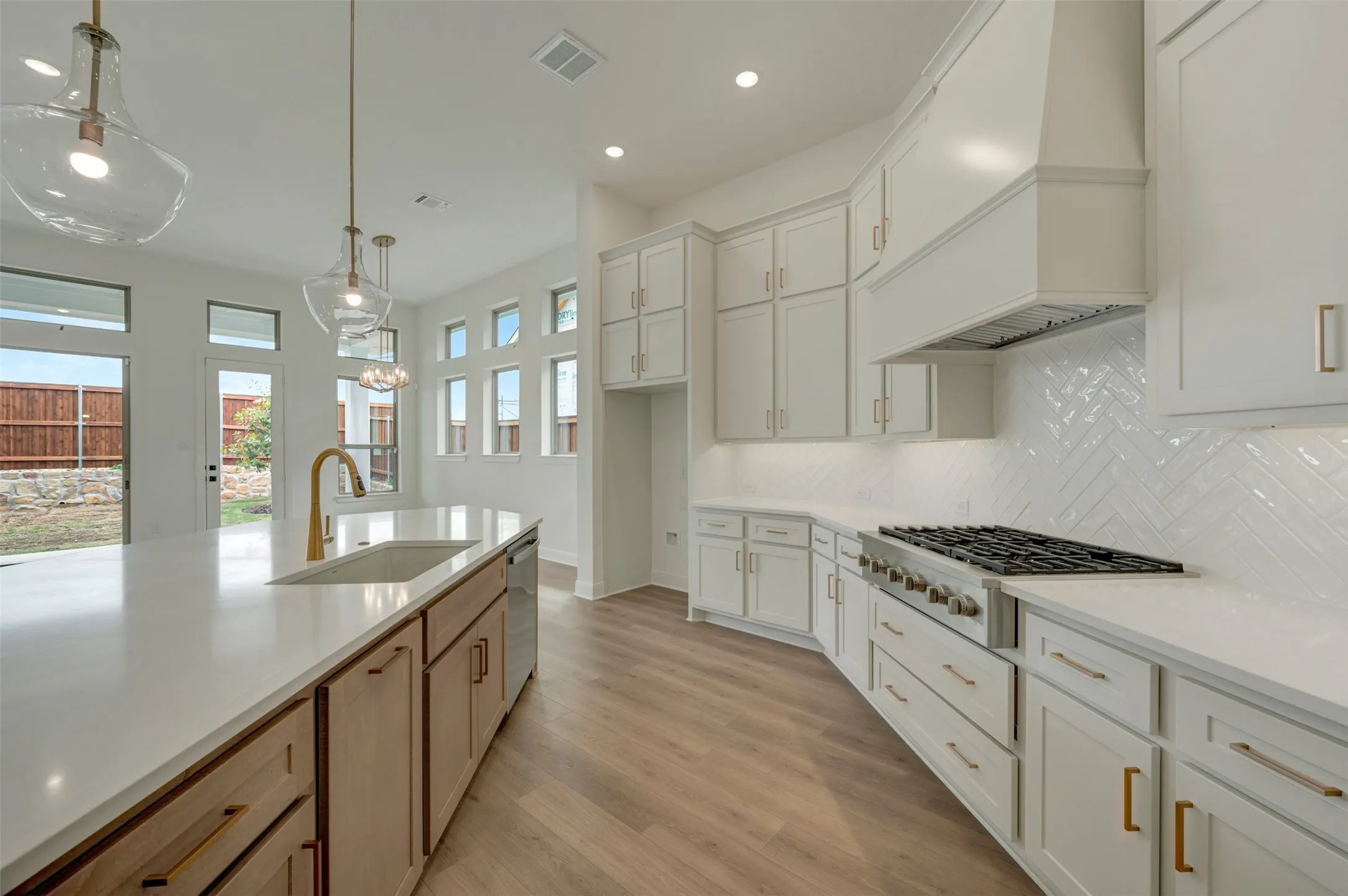 Kitchen featuring an elongated island with integrated sink, white cabinetry with gold-tone hardware, wood-finish flooring, a herringbone tile backsplash, and a stainless steel gas range with a matching hood