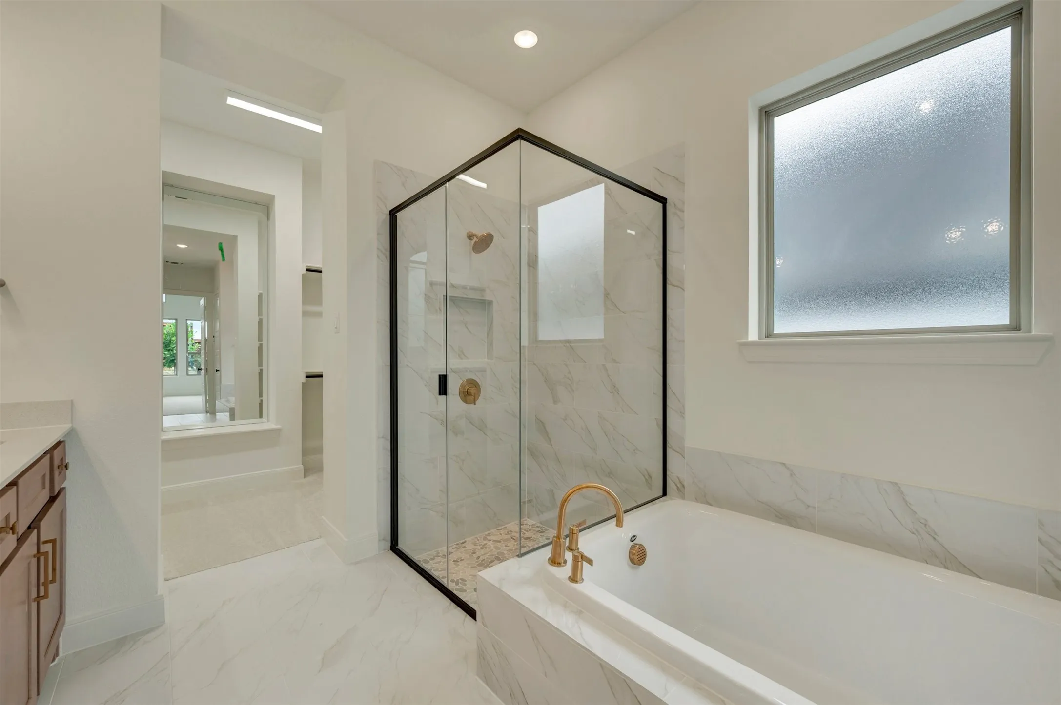 Freestanding bathtub with brass fixtures, glass-enclosed shower with black framing, white tile flooring, and a window with frosted glass