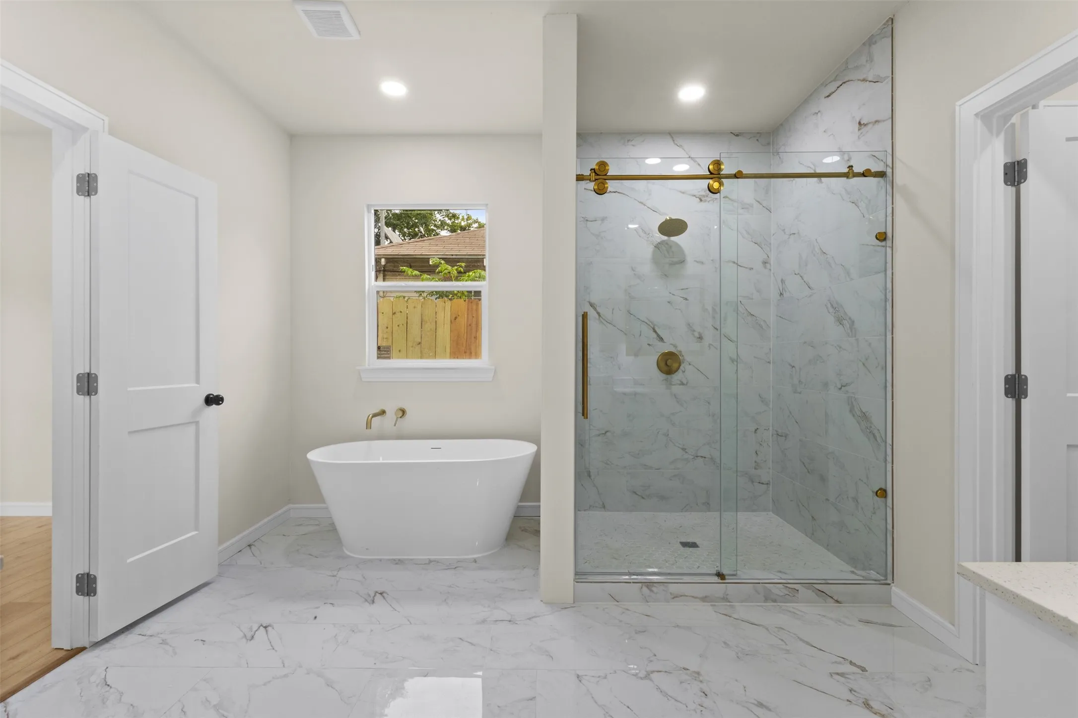 Bathroom featuring a freestanding soaking tub with gold-tone fixtures, a glass-enclosed shower with gold-tone hardware and marble-patterned tile, and large format marble-patterned floor tiles