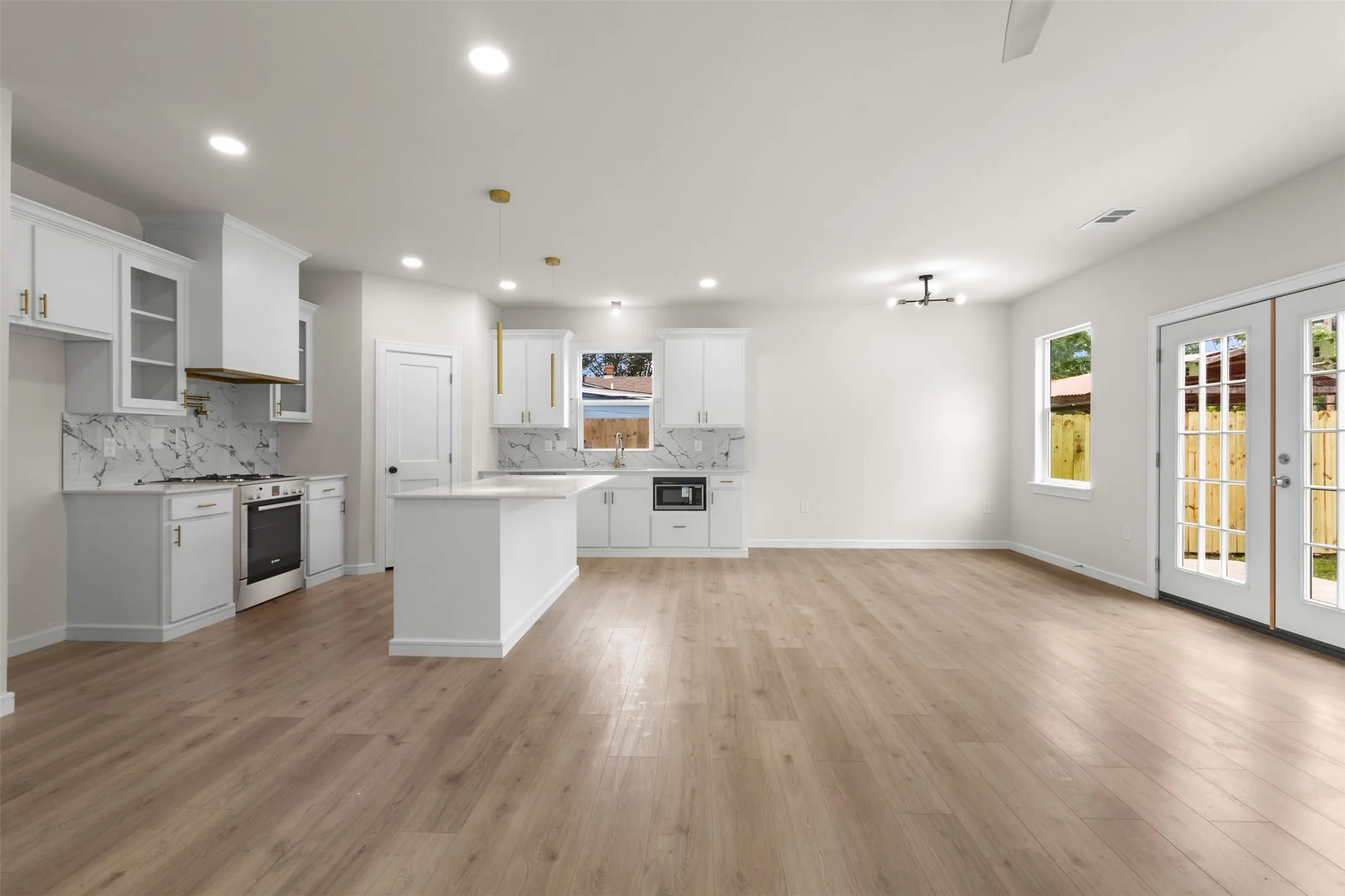 Open-concept living space featuring wood-finish flooring throughout, a modern kitchen with white cabinetry and an island, a gas range with a contemporary range hood, and double French doors to the exterior