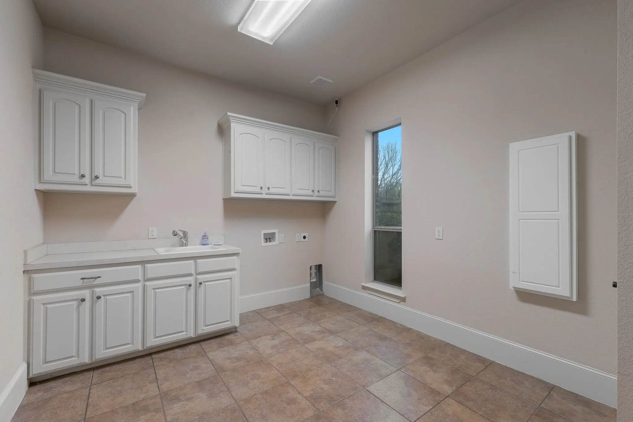 Functional utility room featuring extensive white cabinetry, a built-in sink with a countertop, tiled flooring, and a window providing natural light