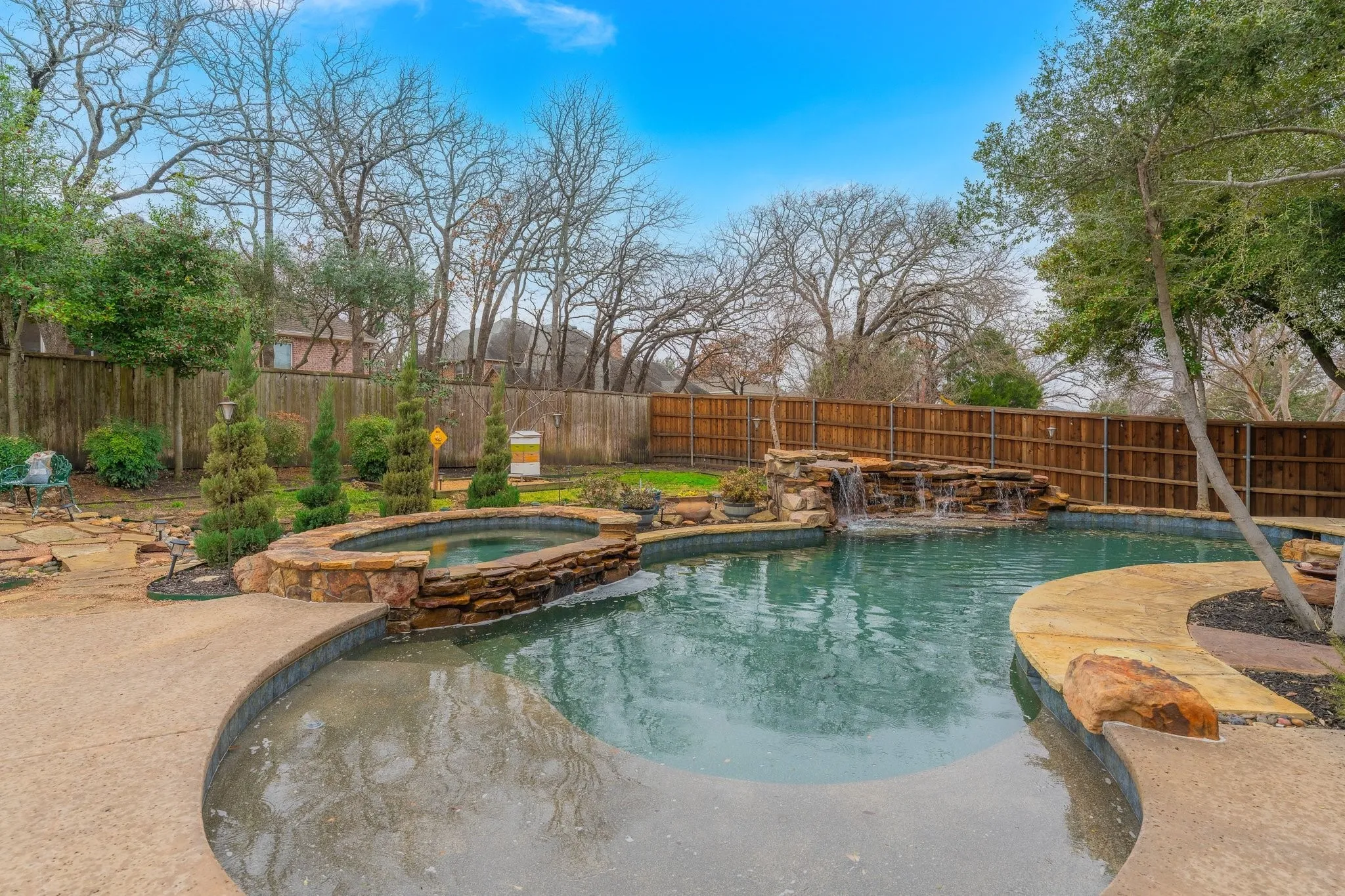 Curved swimming pool featuring a waterfall, integrated spa, stone coping, and surrounding concrete decking