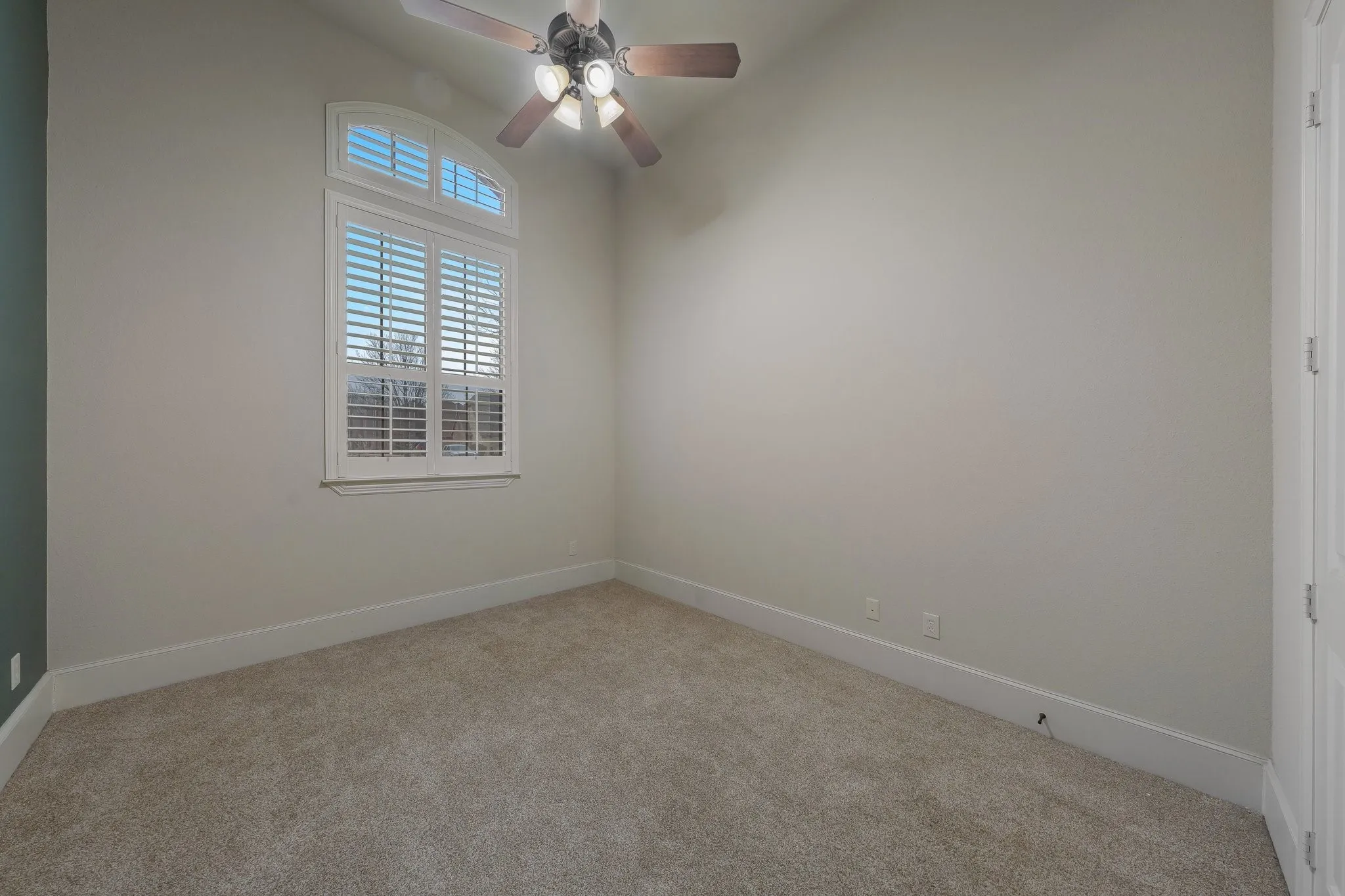 Carpeted room featuring an arched window with white plantation shutters, a ceiling fan with light fixture, and neutral wall tones