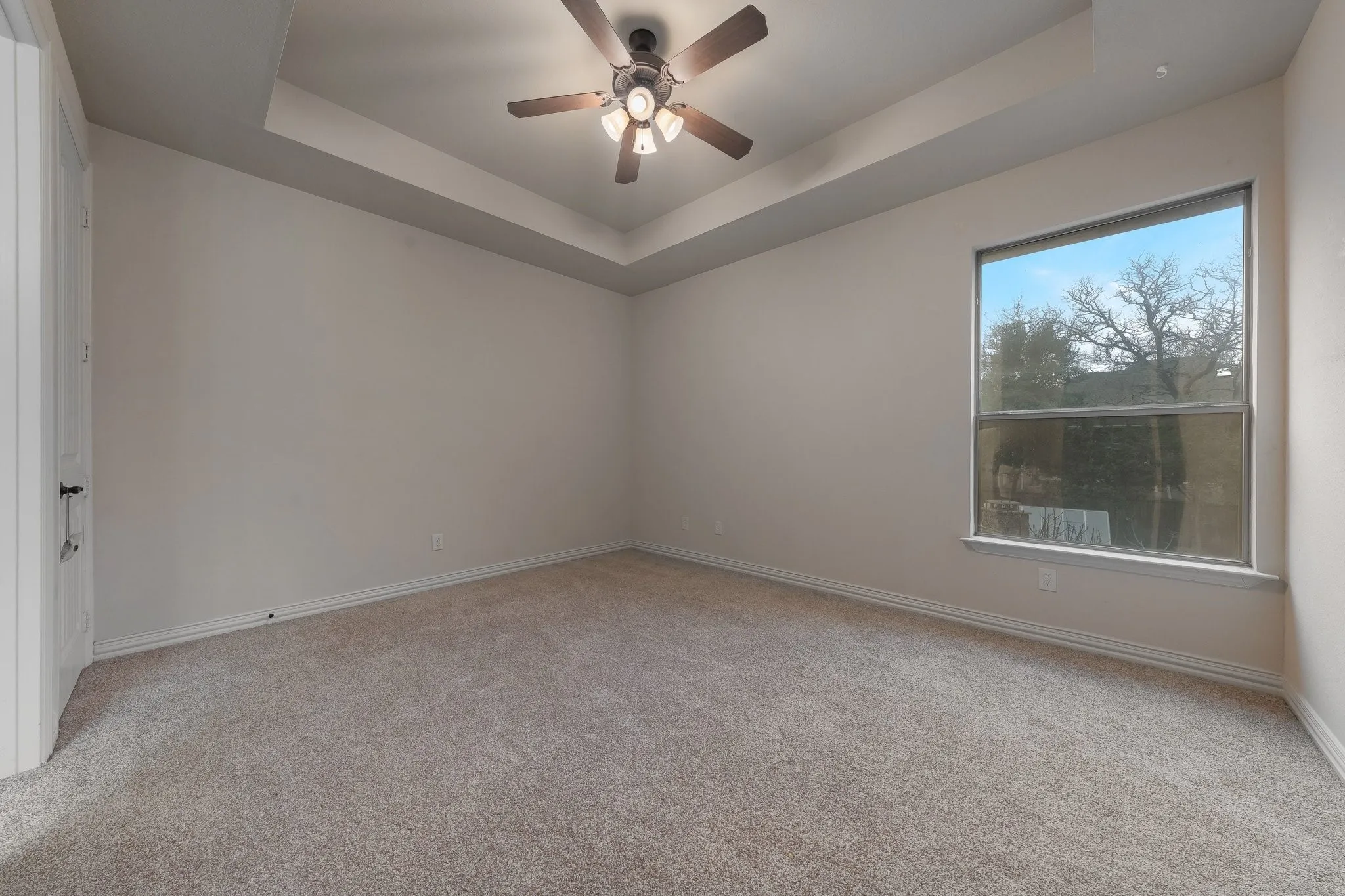 Spacious room featuring neutral carpeting, a tray ceiling with a ceiling fan, and a large window