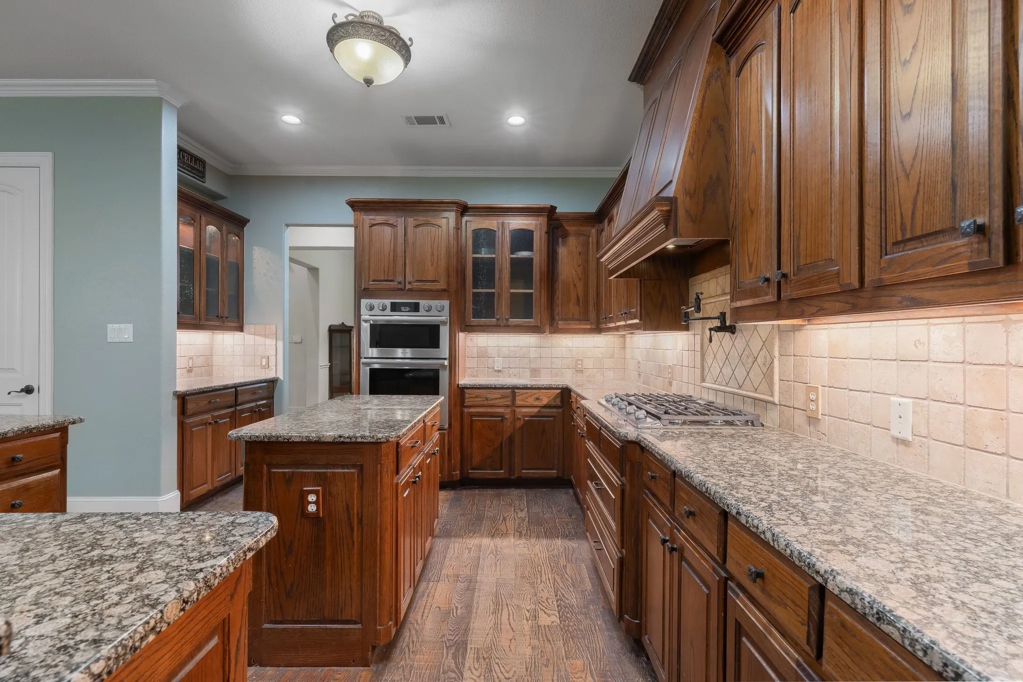 Kitchen featuring wood-finish flooring, custom wood cabinetry, granite countertops, tile backsplash, and stainless steel double ovens