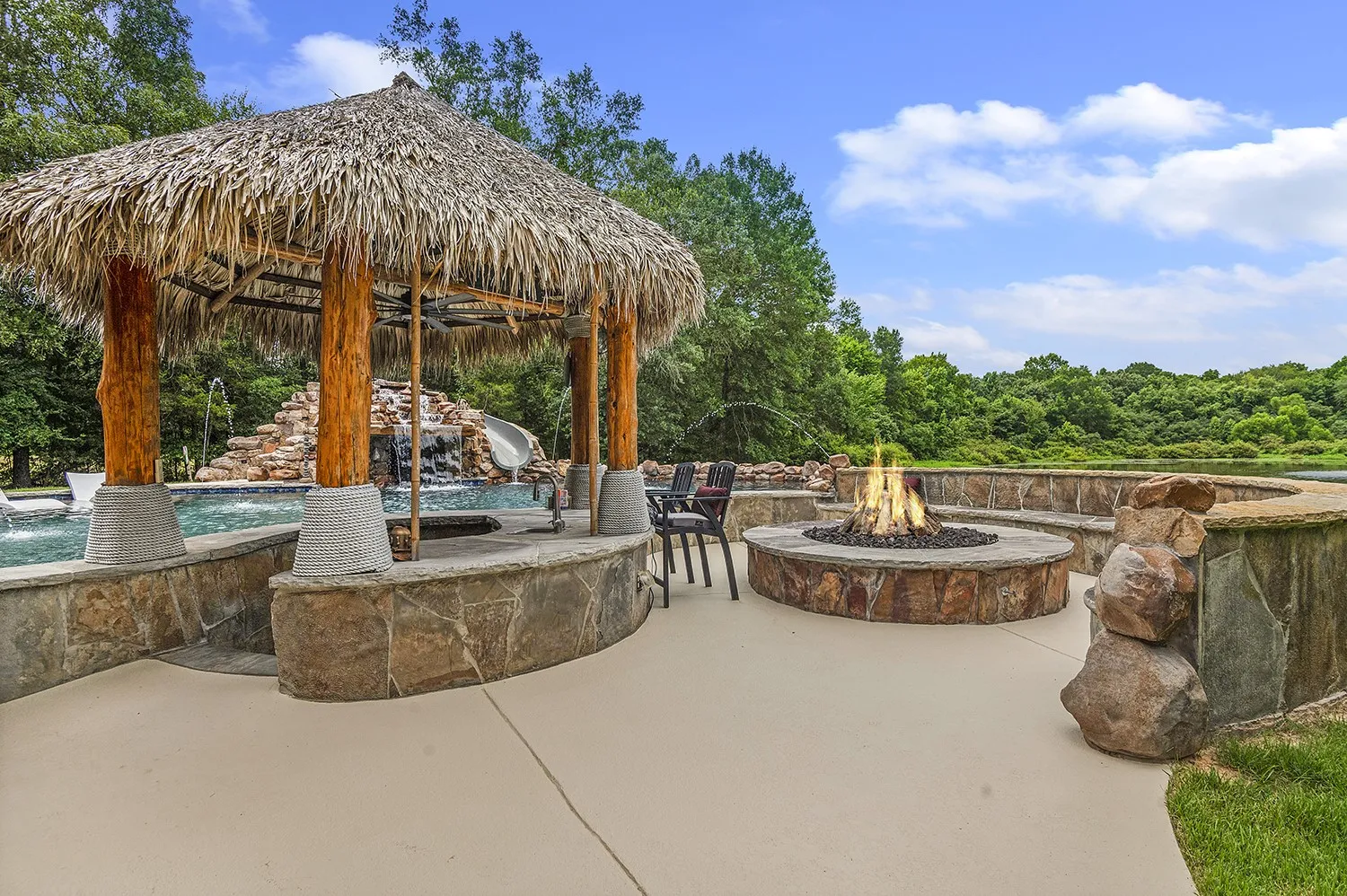 Expansive outdoor living area featuring a thatched-roof cabana, a custom fire pit with stone surround and built-in bench seating, a swimming pool with a rock waterfall and slide, and a surrounding paved patio