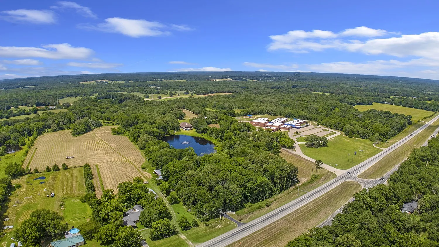 Expansive rural landscape featuring a private pond with fountain, mature trees, open fields, and paved road frontage