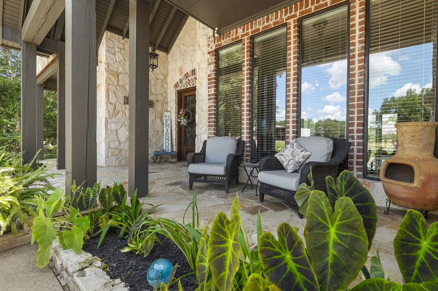 Stone and brick facade featuring a covered patio with flagstone flooring, wood support columns, and lush landscaping