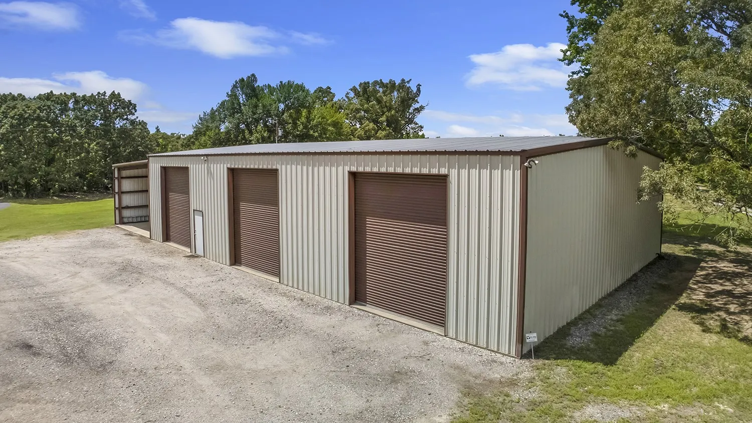 Metal building featuring a corrugated panel exterior, dark brown roll-up doors, and a detached open-sided extension for RV parking