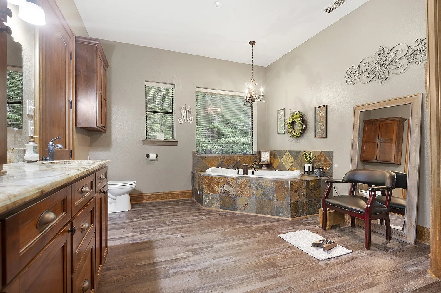 Bathroom featuring a large corner soaking tub with slate tile surround, dual windows with privacy blinds, wood-finish flooring, and a double vanity with stone countertop and wood cabinetry