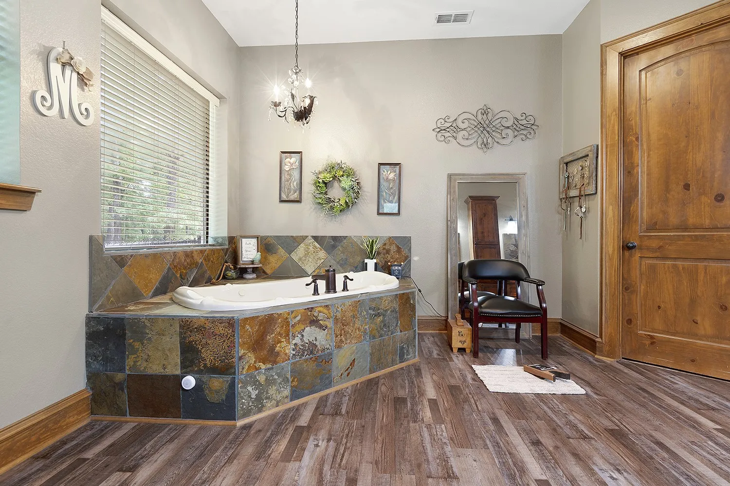 Soaking tub with natural stone tile surround, wood-finish flooring, and a clear-pane window
