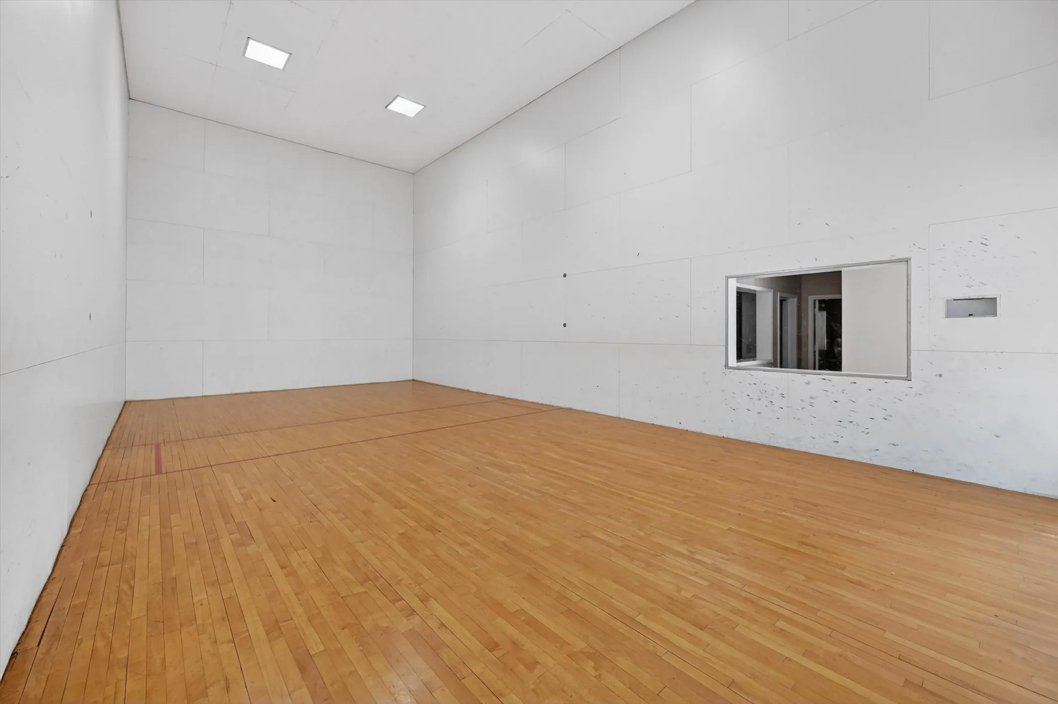 Indoor court space featuring hardwood flooring with court line markings, white paneled walls, recessed ceiling lighting, and an observation window
