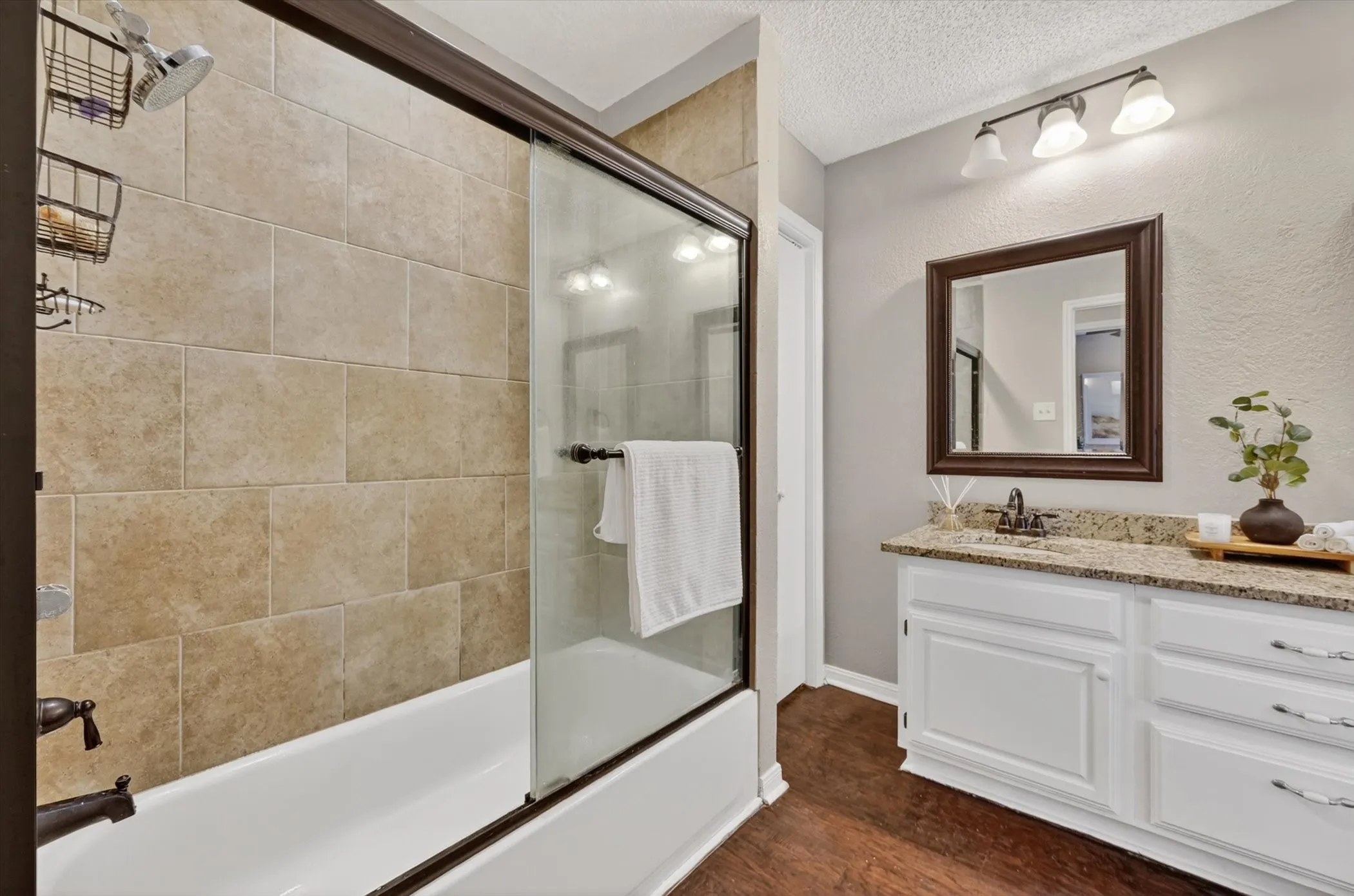 Full bathroom featuring a tub-shower combination with sliding glass doors, large format tile surround, a single vanity with granite countertop, and wood-finish flooring