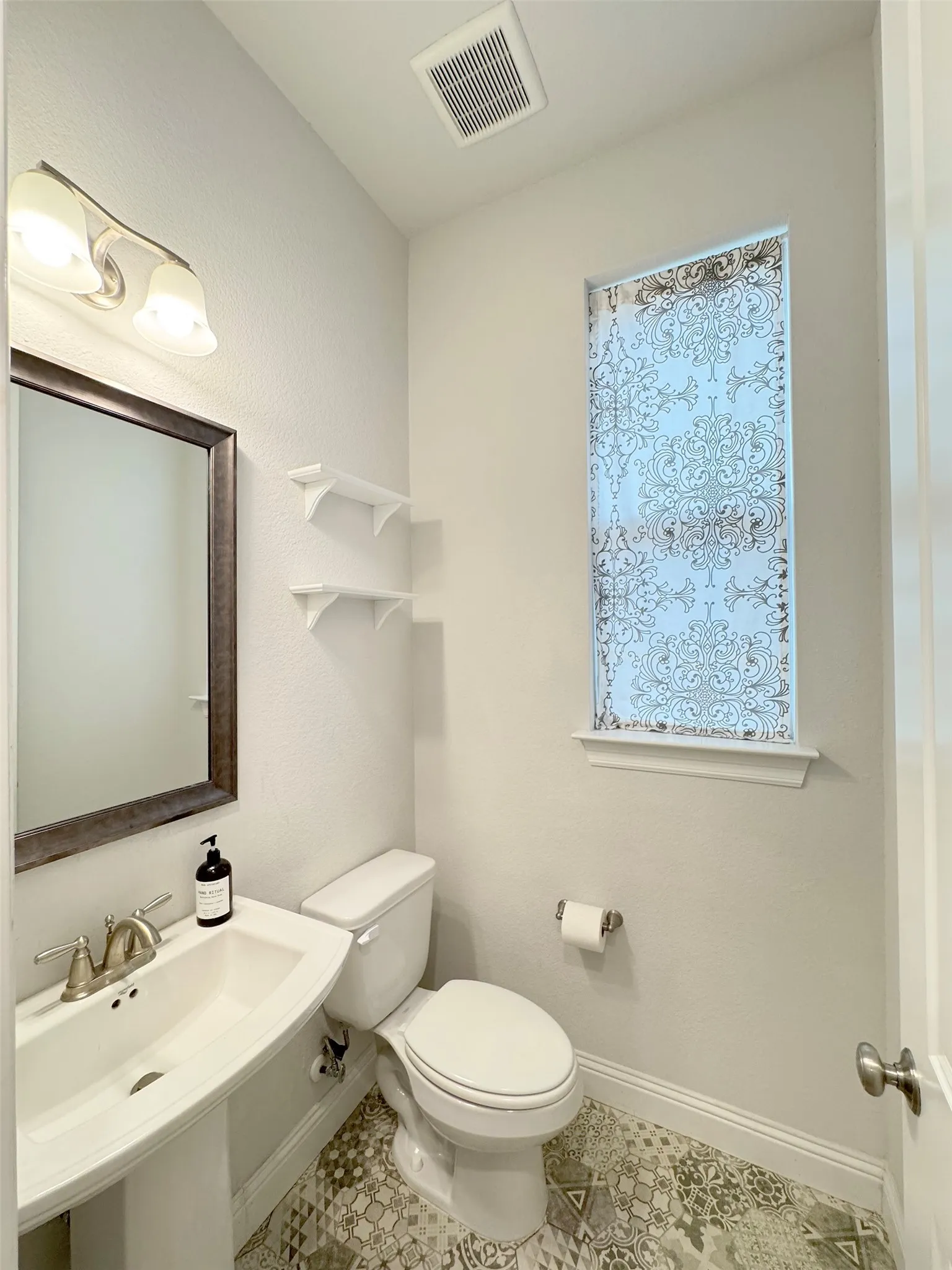 Pedestal sink with brushed nickel faucet, mirror with dark wood-finish frame, and decorative patterned window film