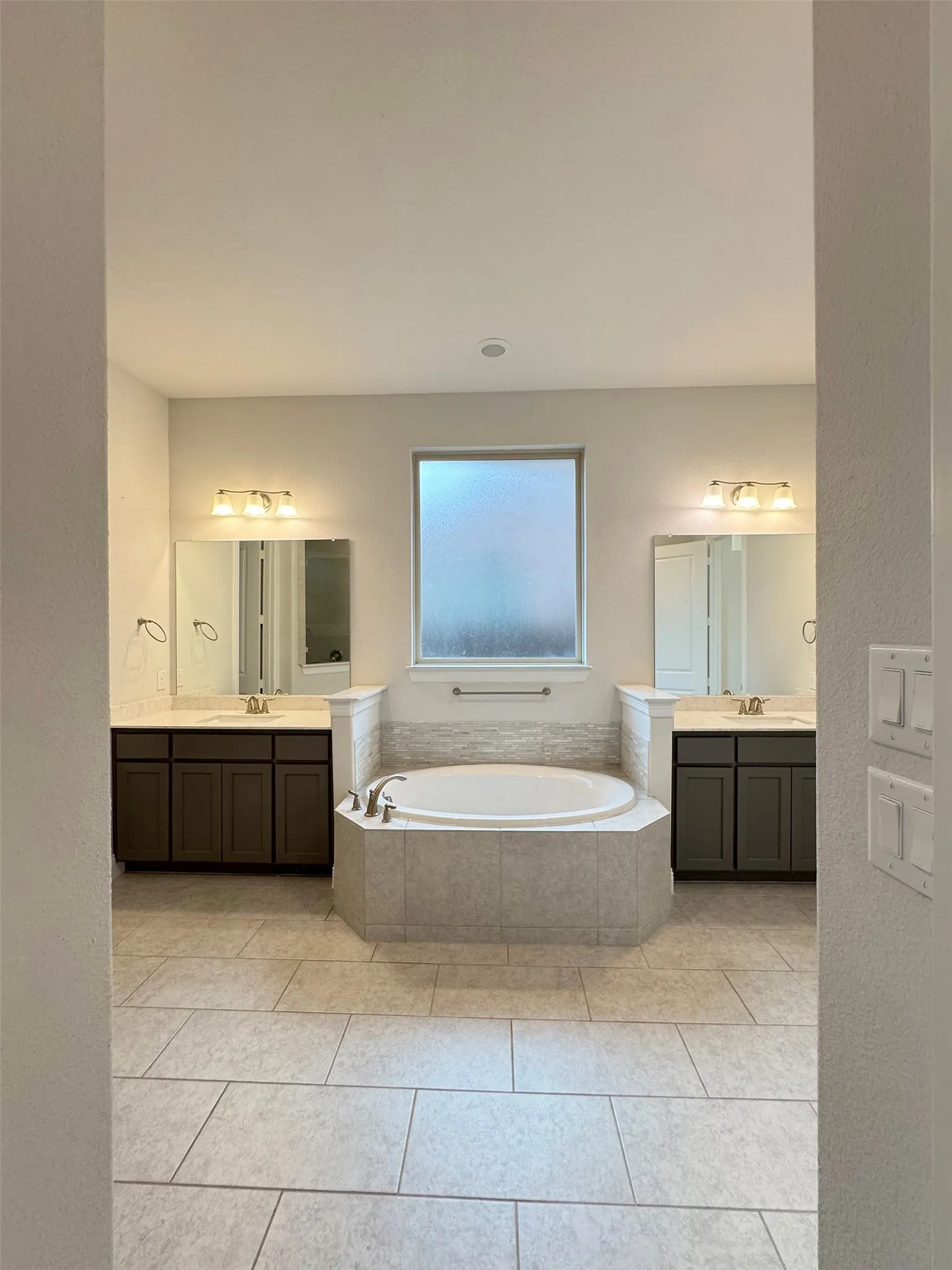 Spacious bathroom featuring a built-in soaking tub with surrounding tile, dual vanity sinks with dark cabinetry, tile flooring, and a frosted window