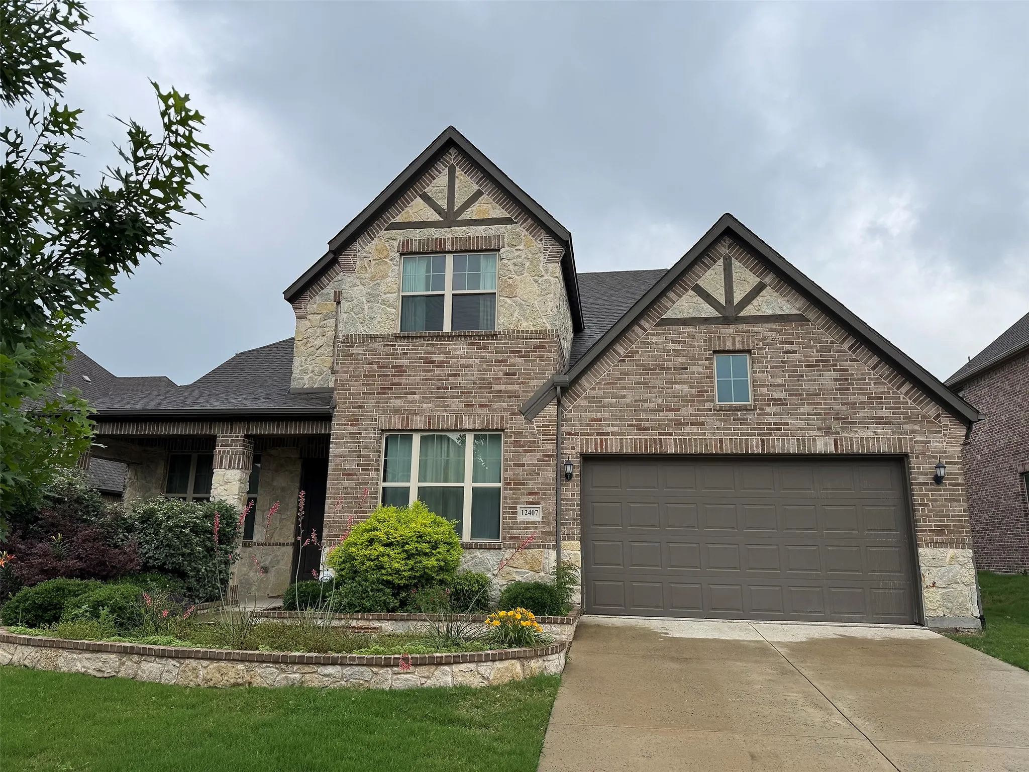 Brick and stone facade featuring a two-car garage, multiple gables, an inviting covered entry, and a decorative stone planter with established landscaping