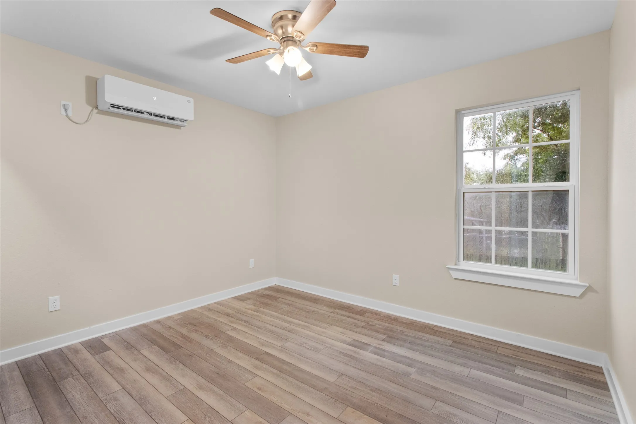 Room featuring wood-finish flooring, a window with grid panes, a ceiling fan with light fixture, and a wall-mounted mini-split HVAC unit