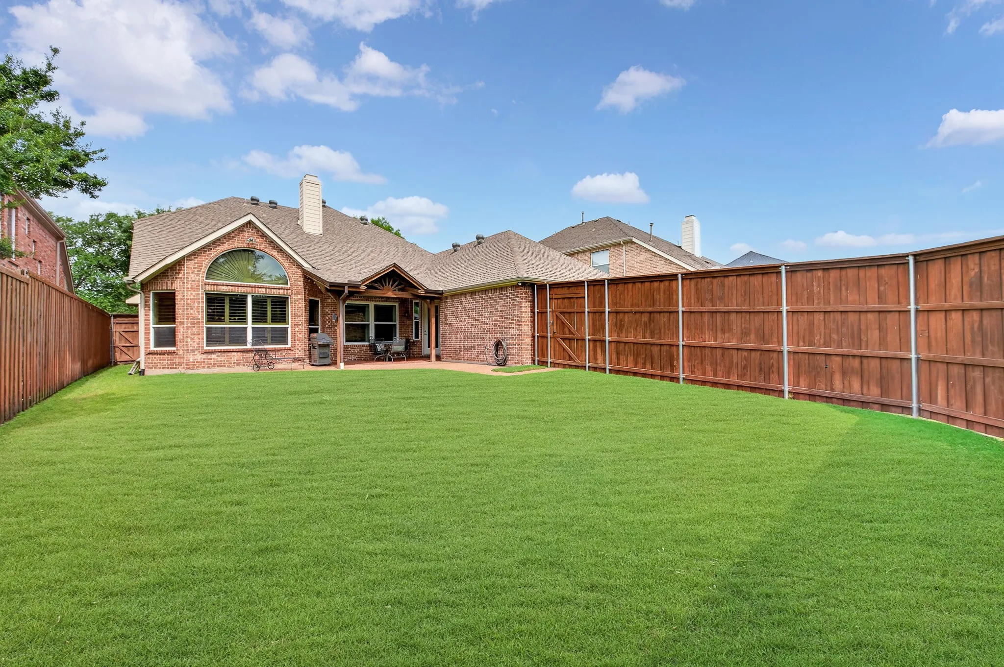 Expansive backyard featuring a manicured lawn, full privacy fencing, brick exterior, covered patio, and a prominent arched window