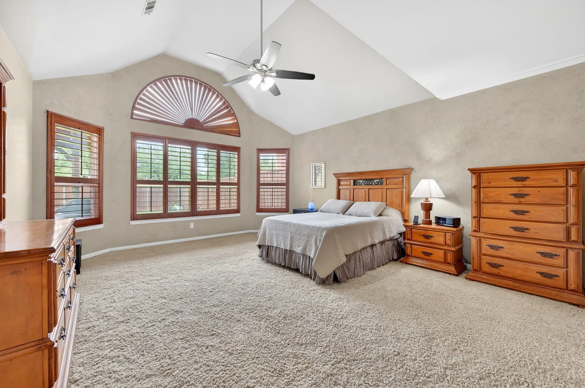 Vaulted ceiling bedroom with a ceiling fan, plush carpet flooring, and a large arched window featuring wooden shutters