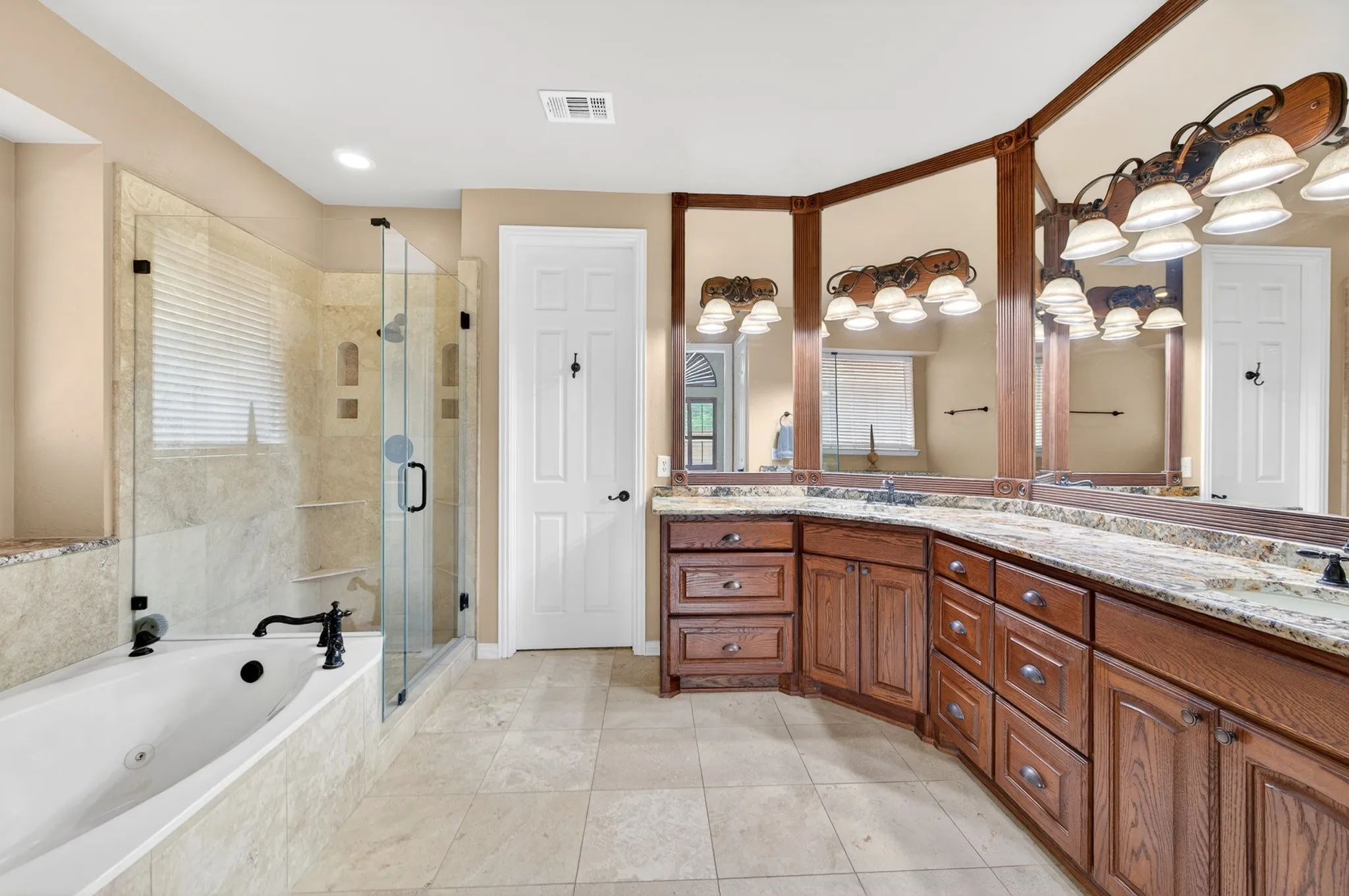 Spacious bathroom featuring a dual vanity with granite countertops, wood cabinetry, an oversized soaking tub, and a glass-enclosed shower with tile surround