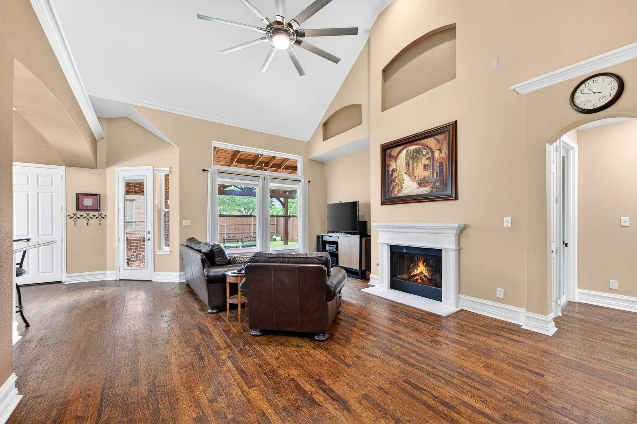 Spacious living area featuring wood-finish flooring, a vaulted ceiling with a ceiling fan, a fireplace with a classic white mantel, and large windows overlooking the backyard
