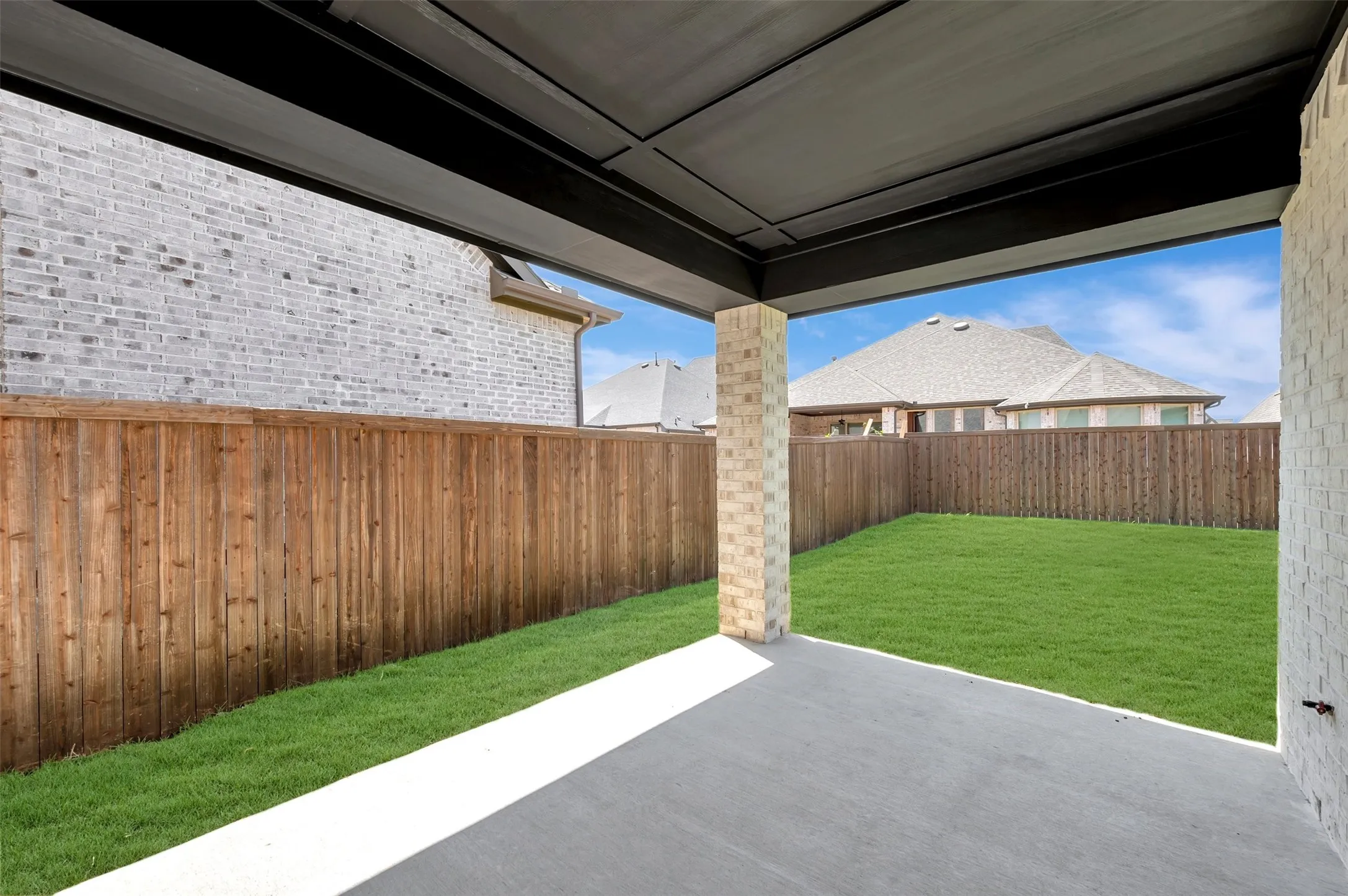 Covered patio with concrete flooring, brick columns, and a grassy yard featuring a wood fence