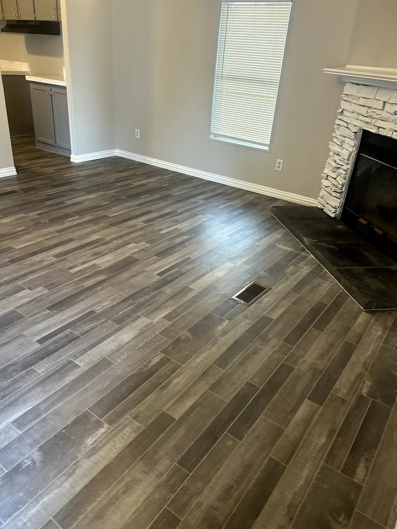 Wood-finish flooring throughout, featuring a stone-clad fireplace with a dark hearth, white baseboards, and a window with blinds