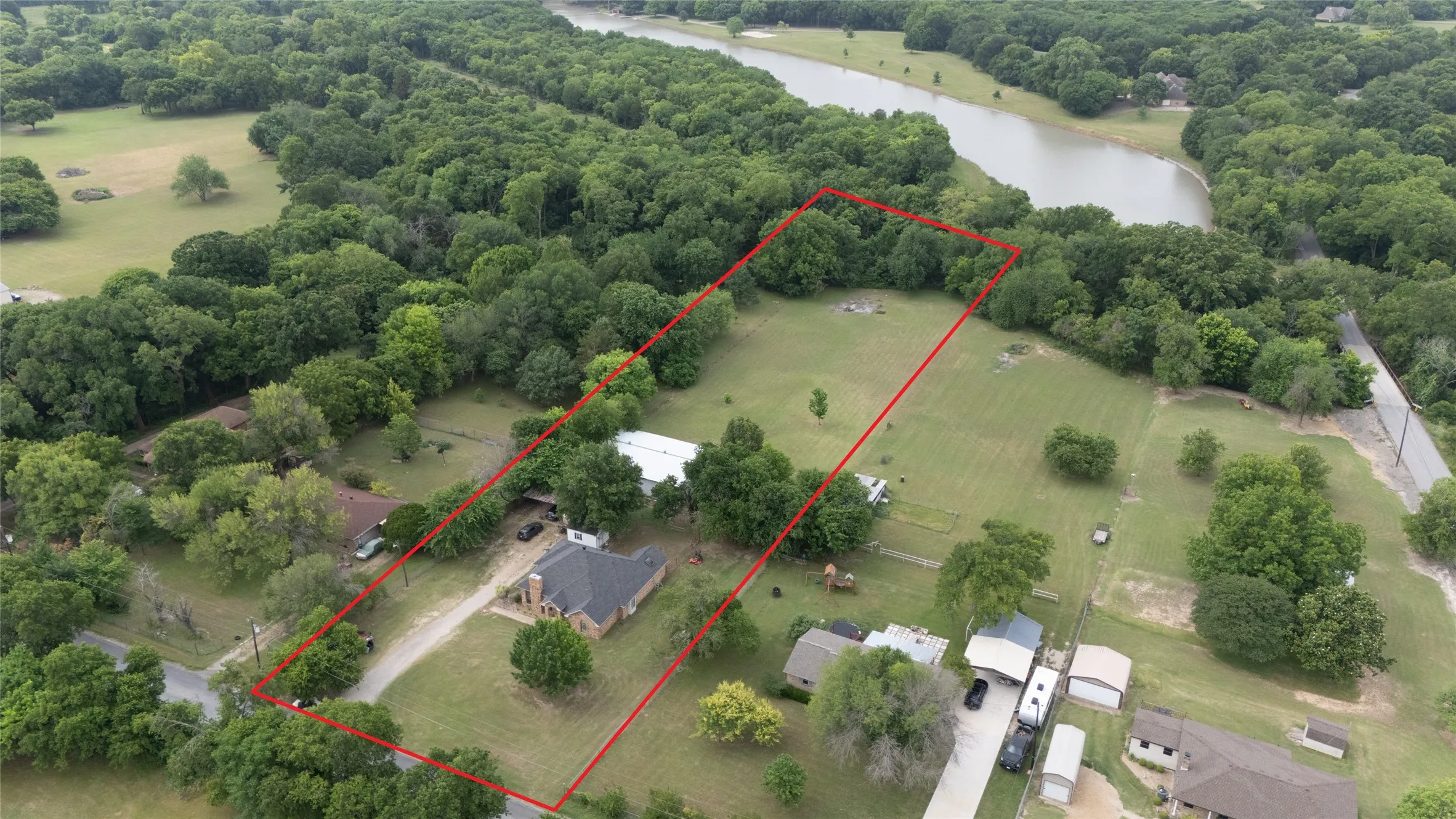 Expansive lot featuring mature trees, a clear water body, and a residential structure with a drive and surrounding green space