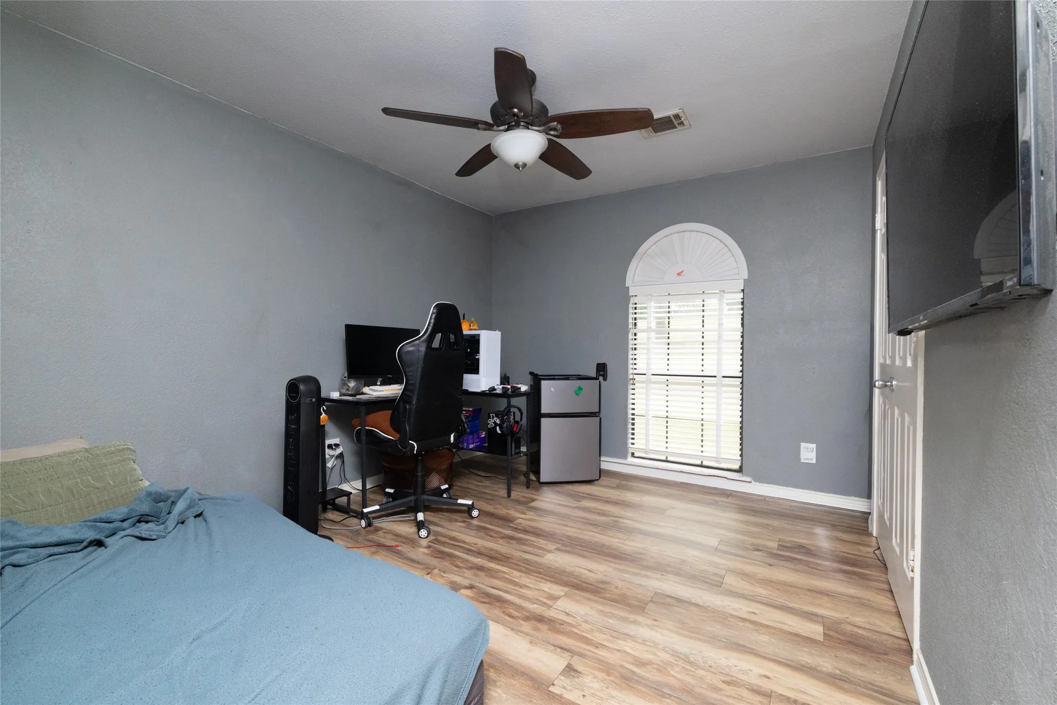 Room featuring wood-look flooring, a ceiling fan with integrated lighting, and a white arched window with blinds