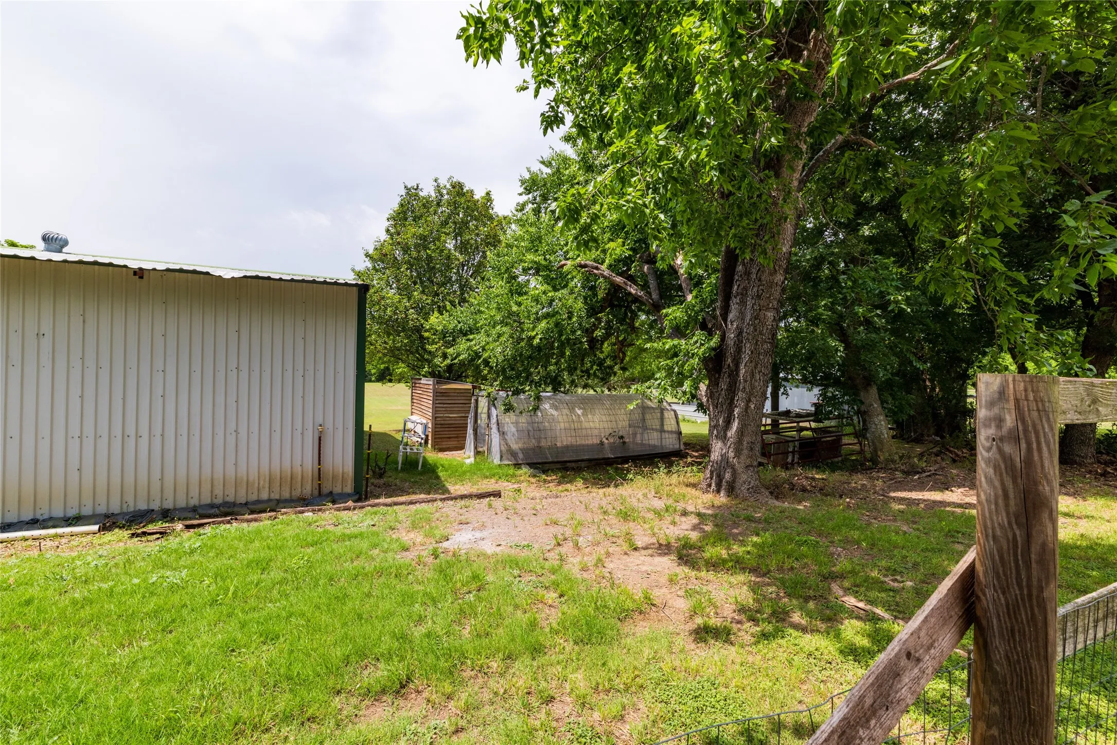 Detached insulated, metal-sided barn with a sloped metal roof, mature trees, and a partially visible fence line, chicken coop and greenhuse