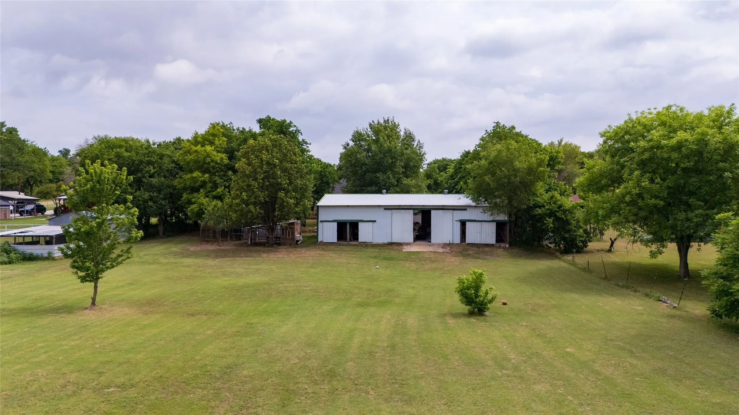Expansive grassy parcel featuring a white barn with multiple bays, a metal roof, and surrounding mature trees