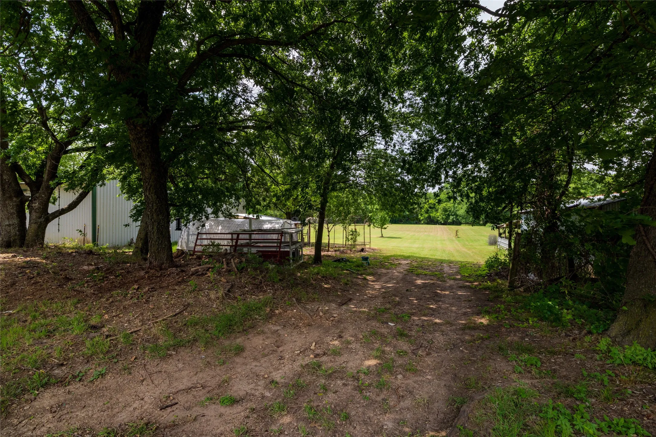 Sprawling agricultural property featuring a dirt access road, extensive open fields, mature shade trees, and a white corrugated metal outbuilding