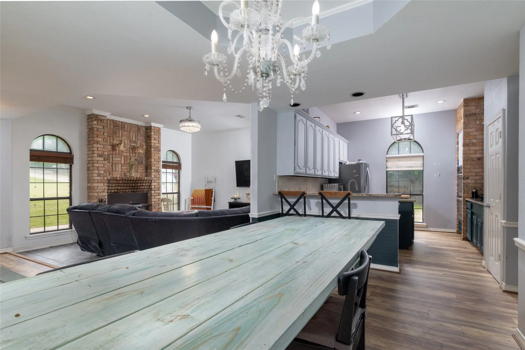 Open-concept dining area featuring wood-look flooring, al chandelier, and direct sightlines to the living space with a brick fireplace and arched windows