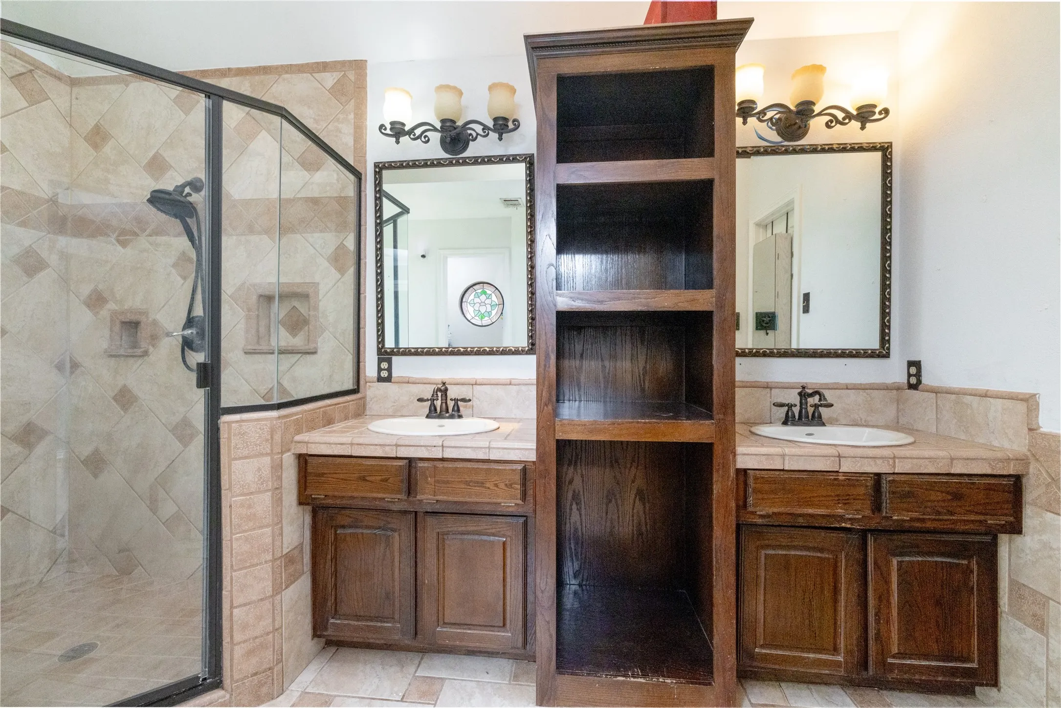 Bathroom featuring a glass-enclosed shower with tile surround, dual wood-finish vanities with tile countertops, and two framed wall mirrors