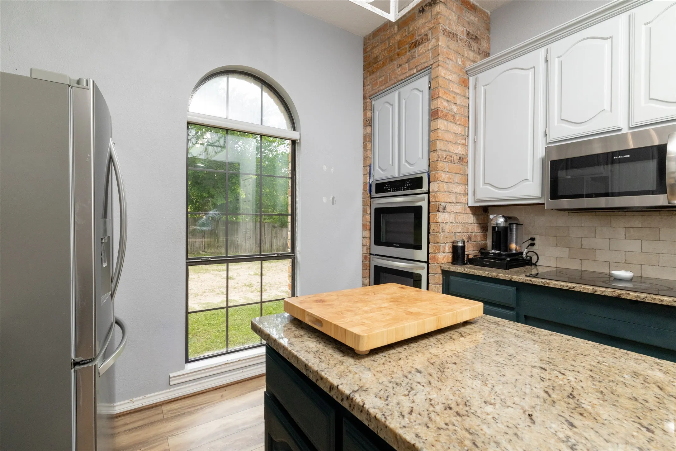 Kitchen featuring a granite countertop island, stainless steel double wall ovens, exposed brick accent, subway tile backsplash, and an arched window