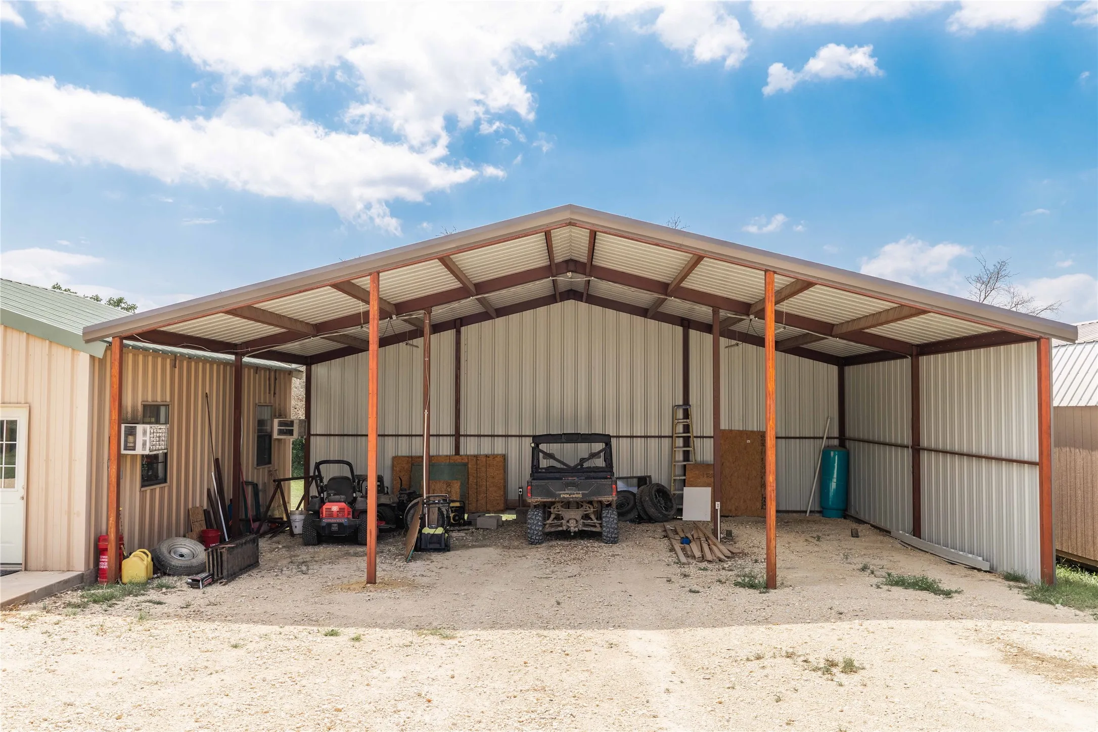 Open-air metal carport with a gravel base, supported by metal posts and a metal roof