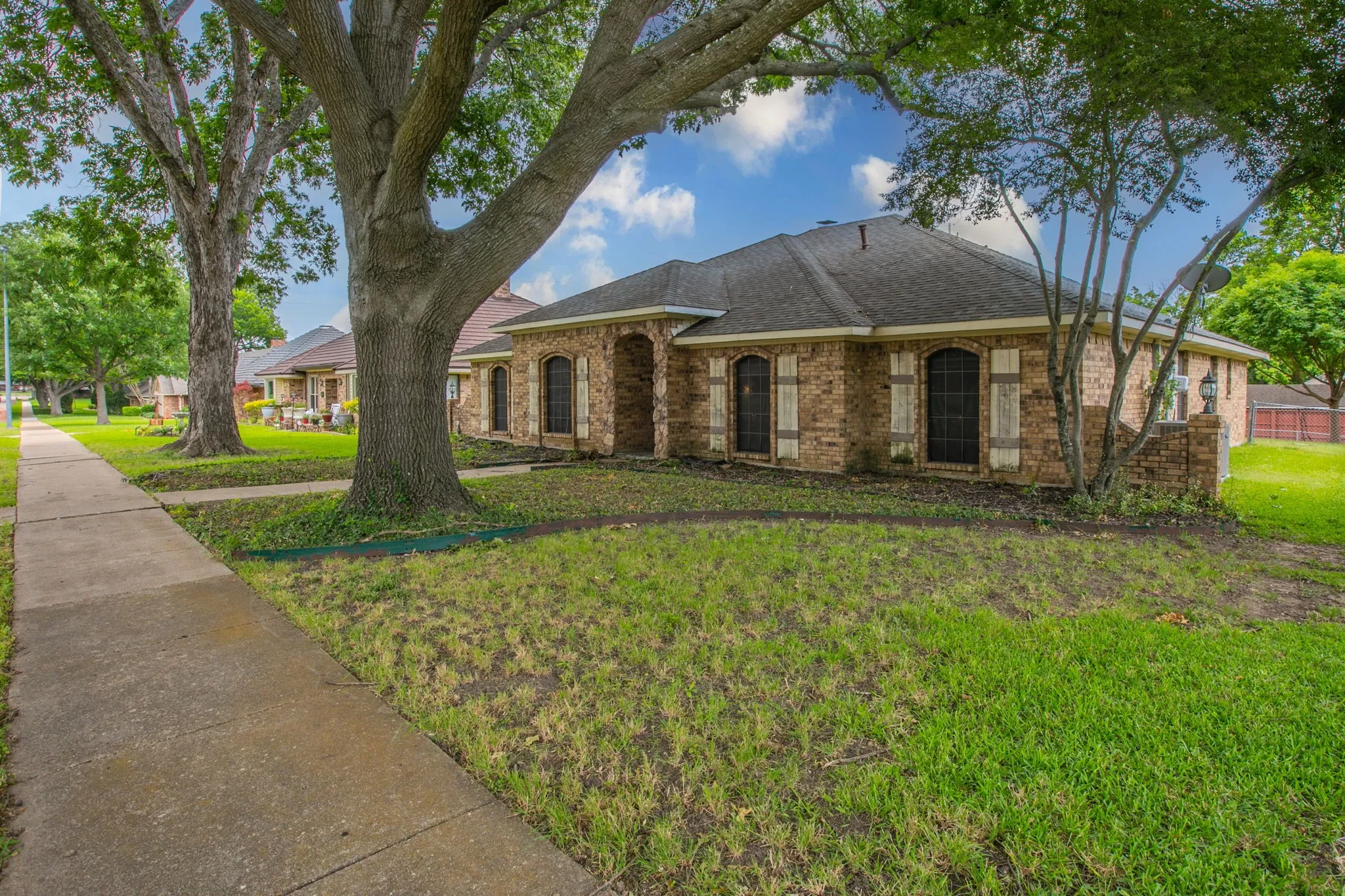 Brick exterior residence featuring arched windows with decorative shutters, a dark shingle roof, and mature trees providing natural shade